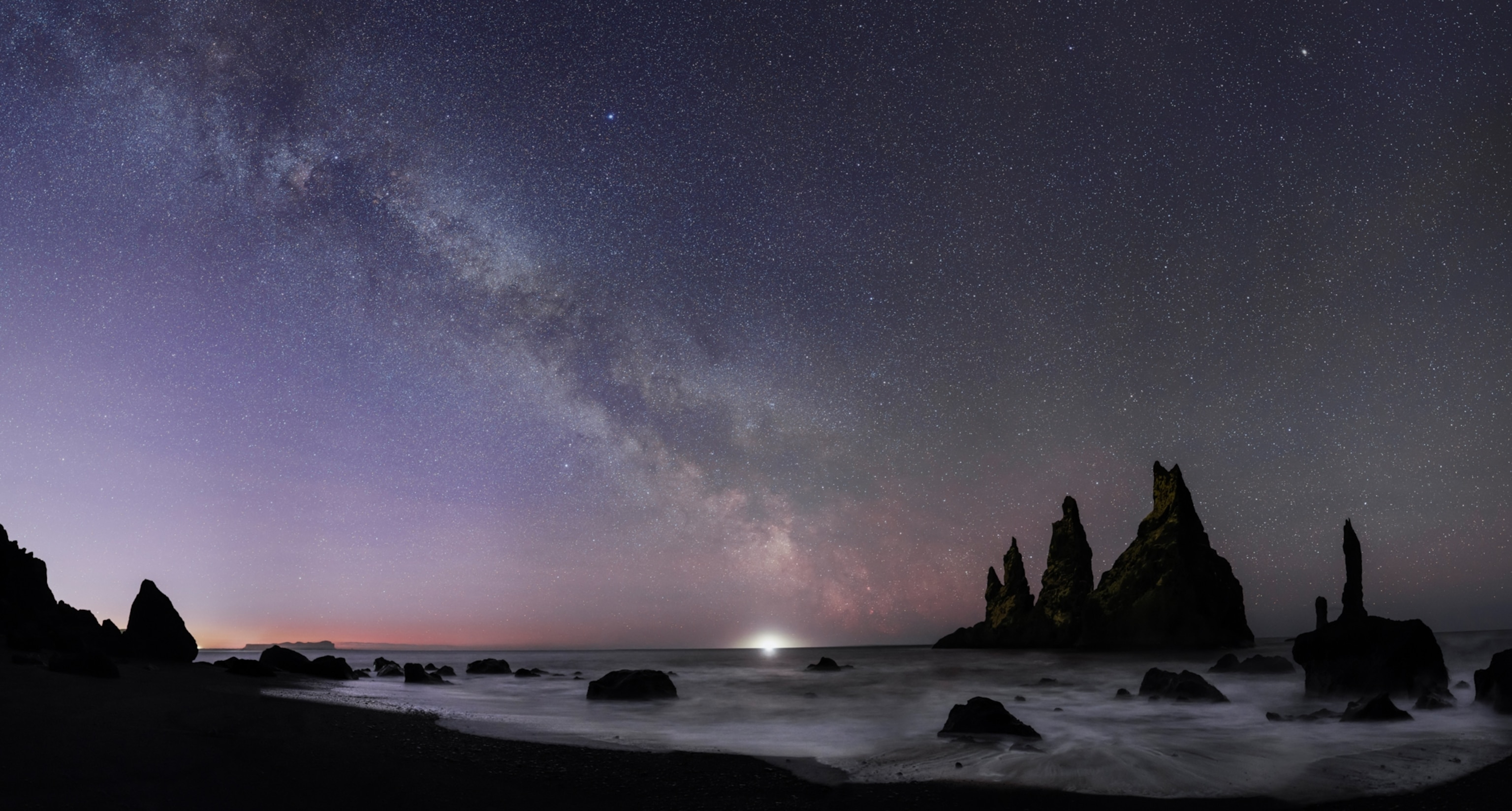 Starry night over a rocky beach with dark sea stacks silhouetted against the sky. The Milky Way stretches across the night sky.