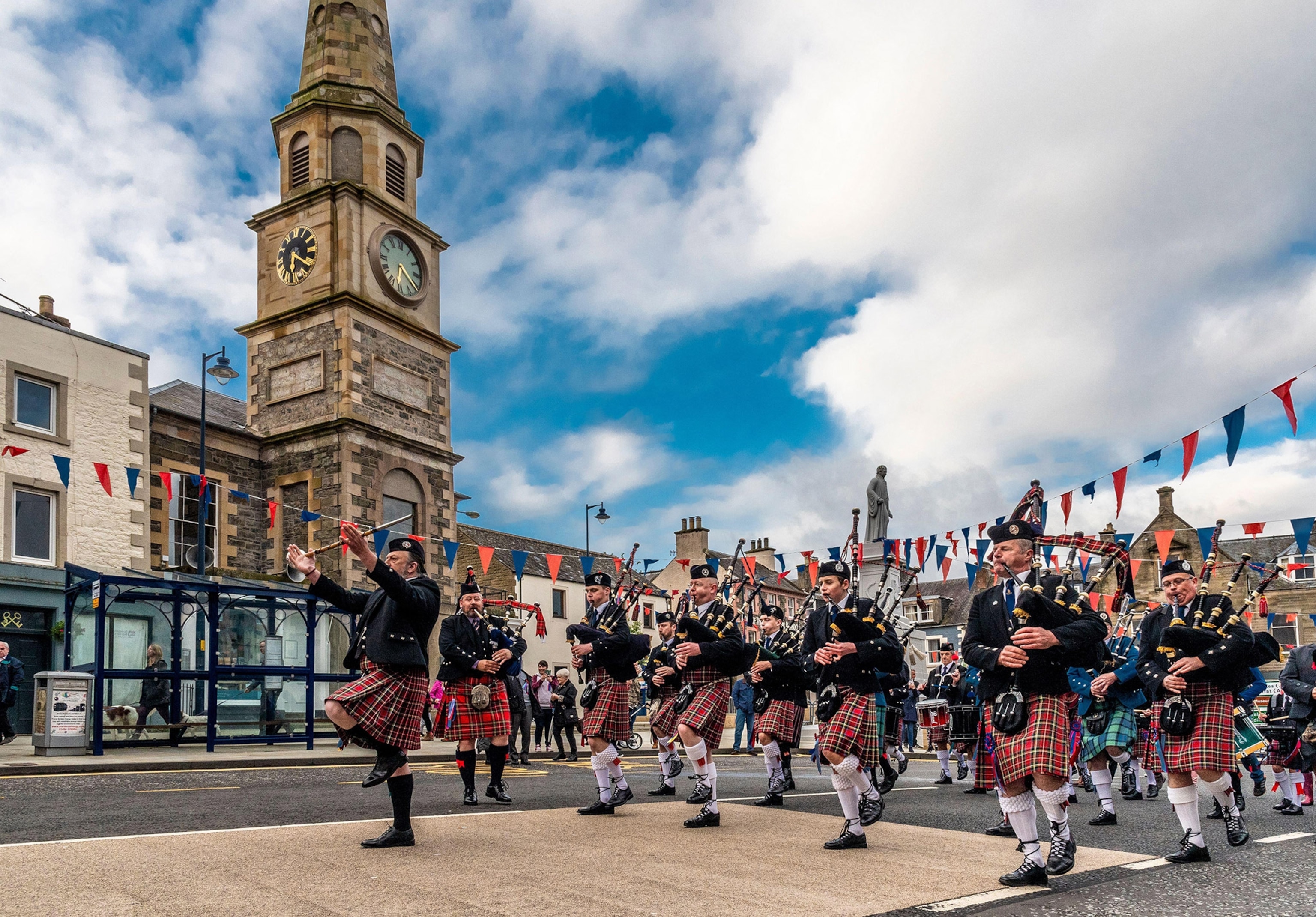 Parade procession featuring people in tartan kilts, some playing bagpipes