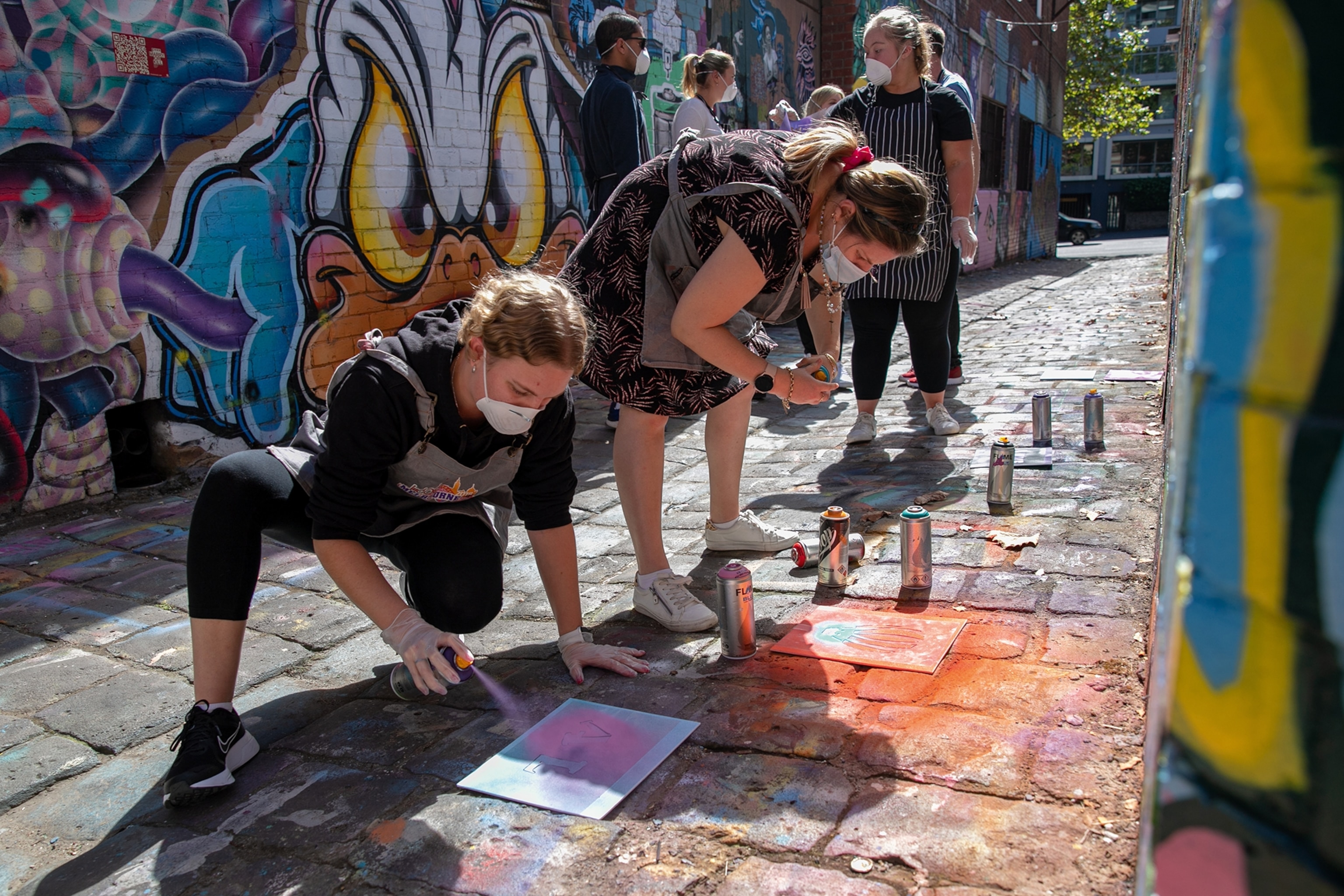 Two women make spray-paint art in front of a wall with graffiti on.