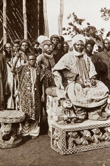 Picture of black and white historical photo of a man in tribal attire sitting on throne and white person next to him on elaborate chair.