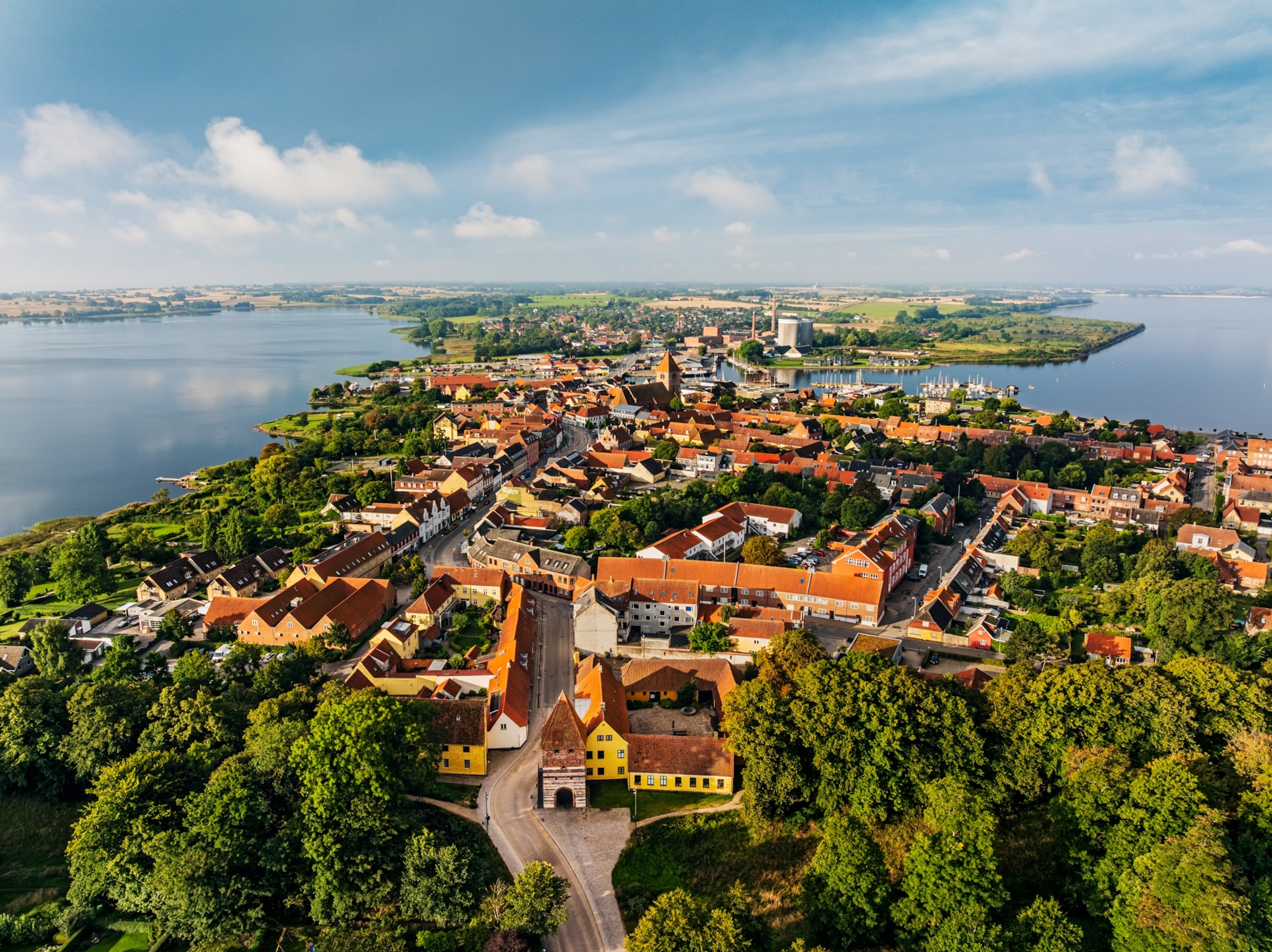 Panoramic aerial drone photograph of the historic market town of Stege on the island of Møn, Denmark. The image shows the town's traditional red-tiled rooftops, winding streets, medieval architecture and waterfront harbour. The historic Mølleporten town gate is visible in the foreground, while Stege Church, the marina and surrounding coastal countryside stretch into the distance beneath a bright summer sky.