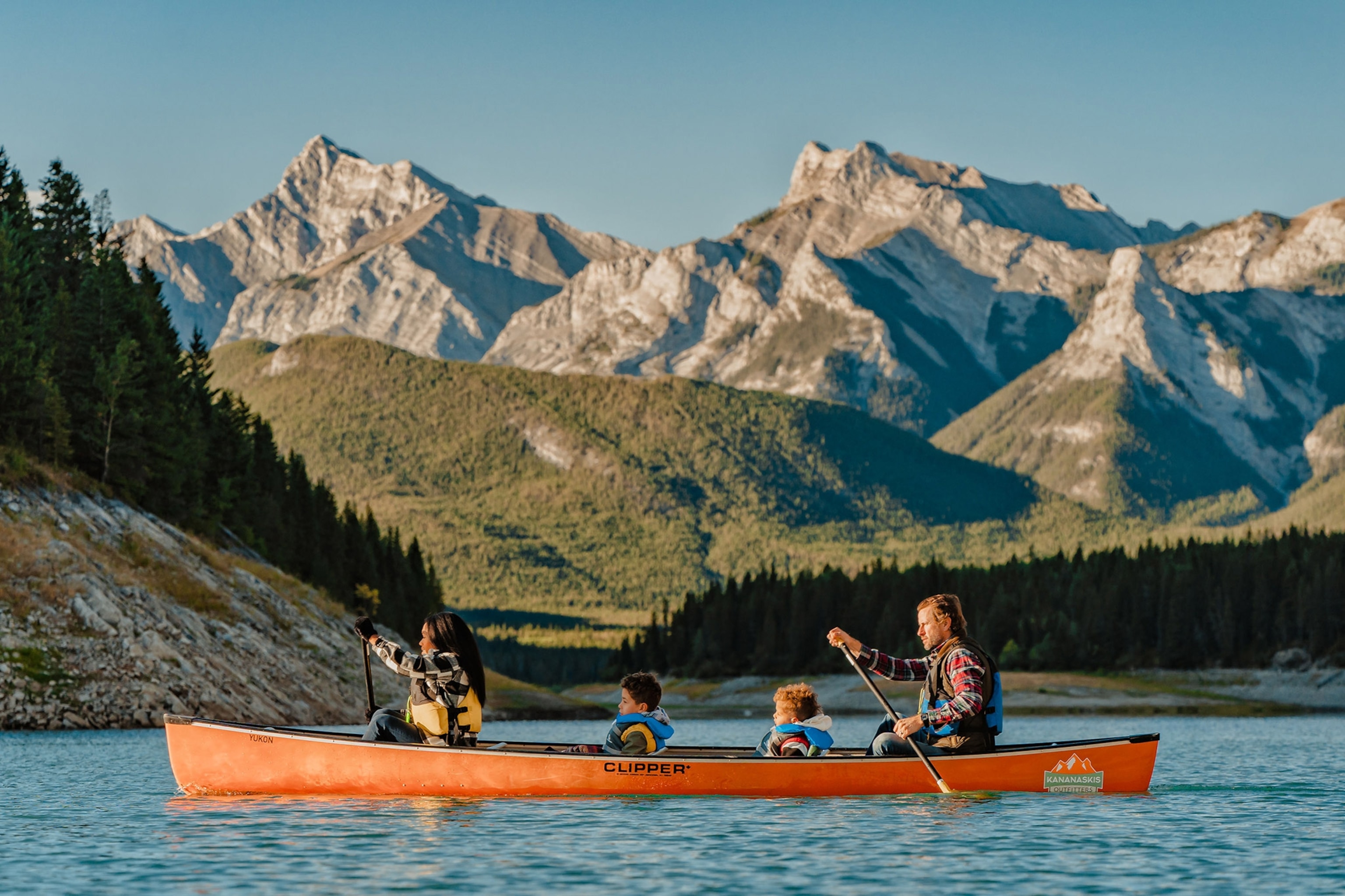 A group of two adults and two children in an orange canoe on a blue lake with mountains behind and a blue sky above