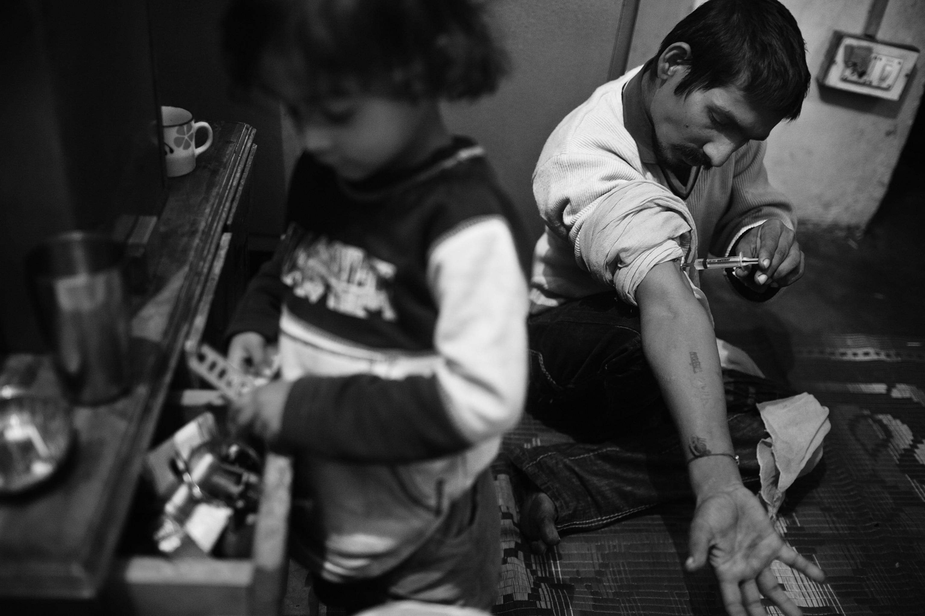 A young girl, left, searches for something in a cupboard drawer, while her father’s friend, Pakori, injects drugs.