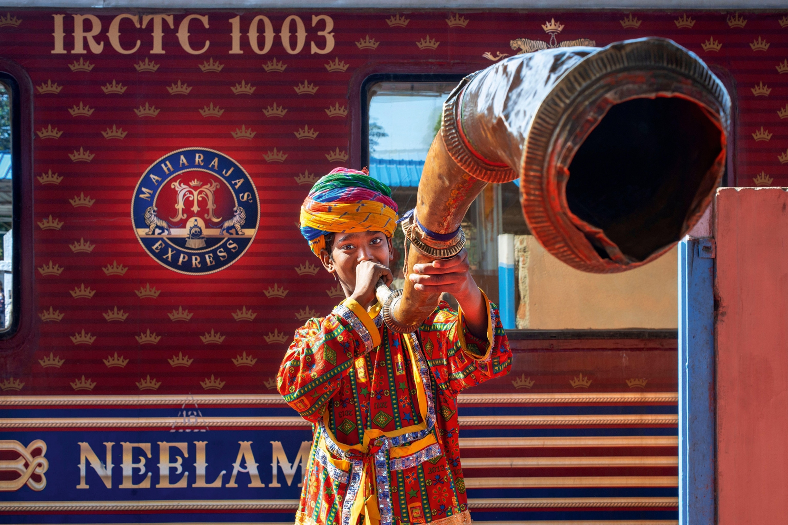 A young, local boy wearing a sequined tunic and turban, blowing a horn in front of a train.