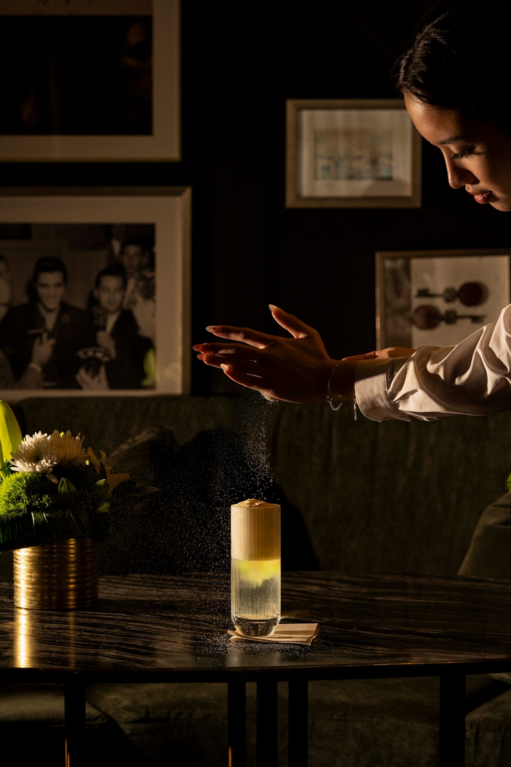 A moody bar table with a long drink as a waitress sieves a fine powder on top.