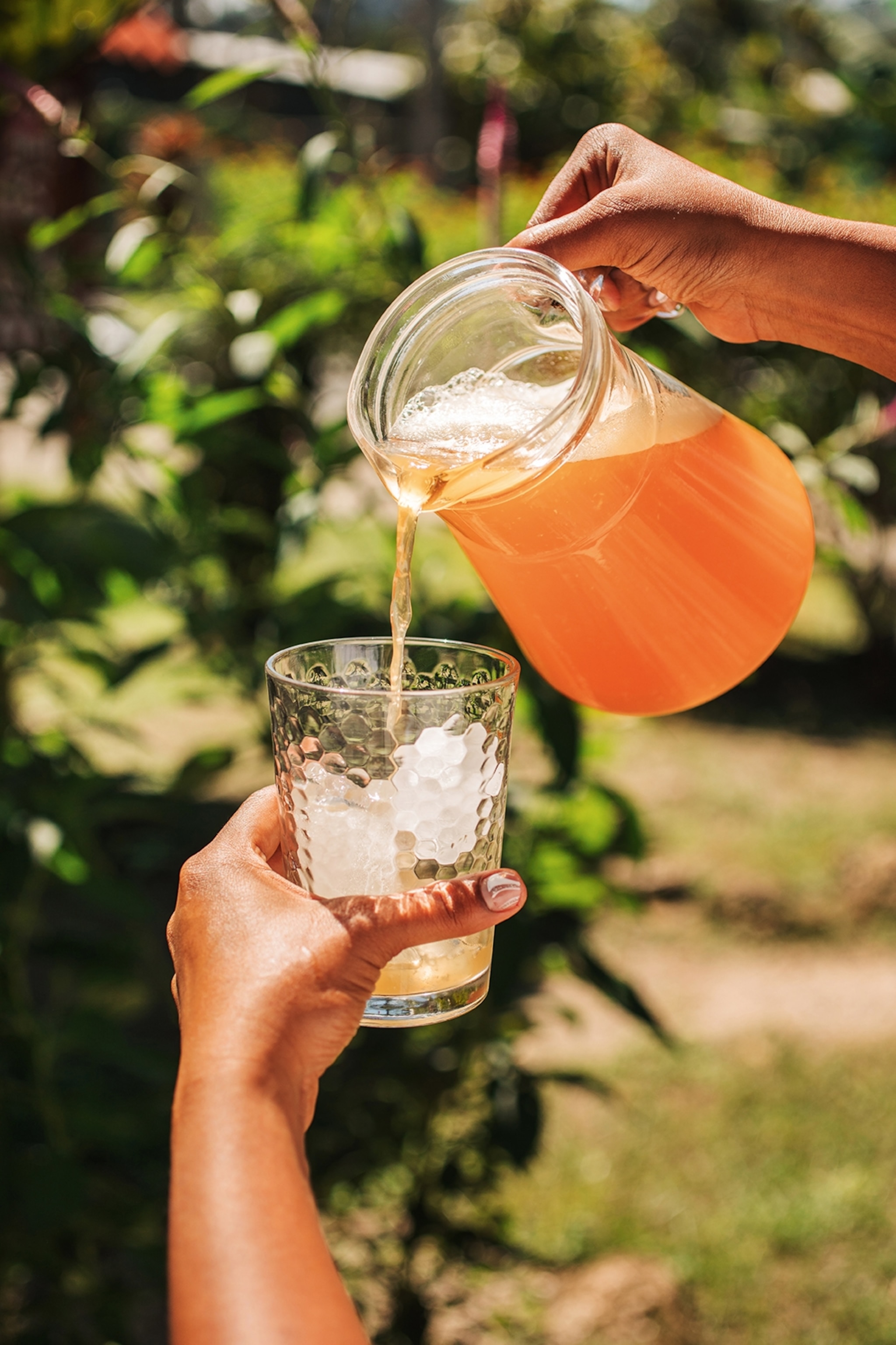 A person pouring an orange lemonade from a jug into a glass with ice.