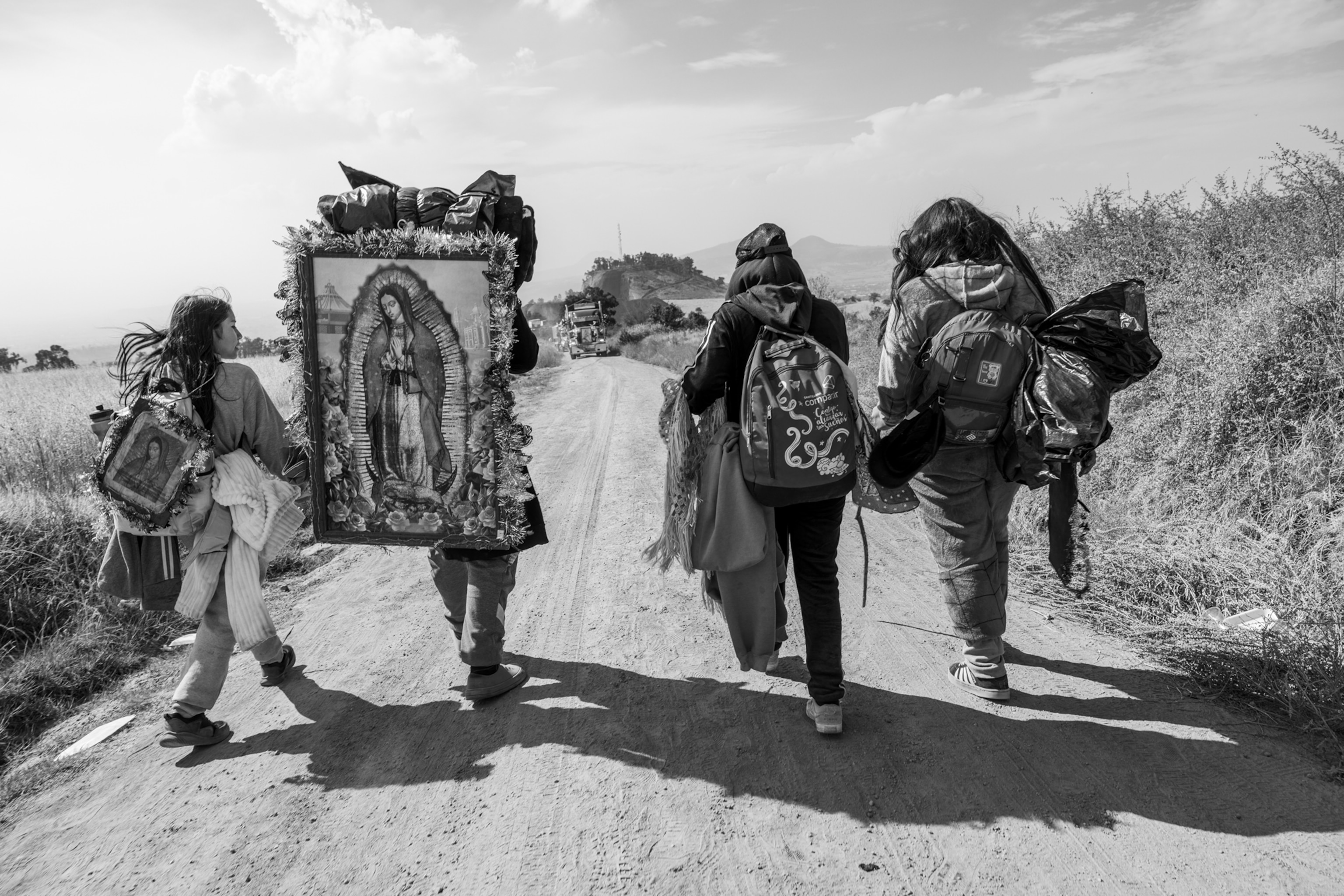 A group of people walk in a line down a dirt road. They have backpacks and a painting of a woman on their backs.