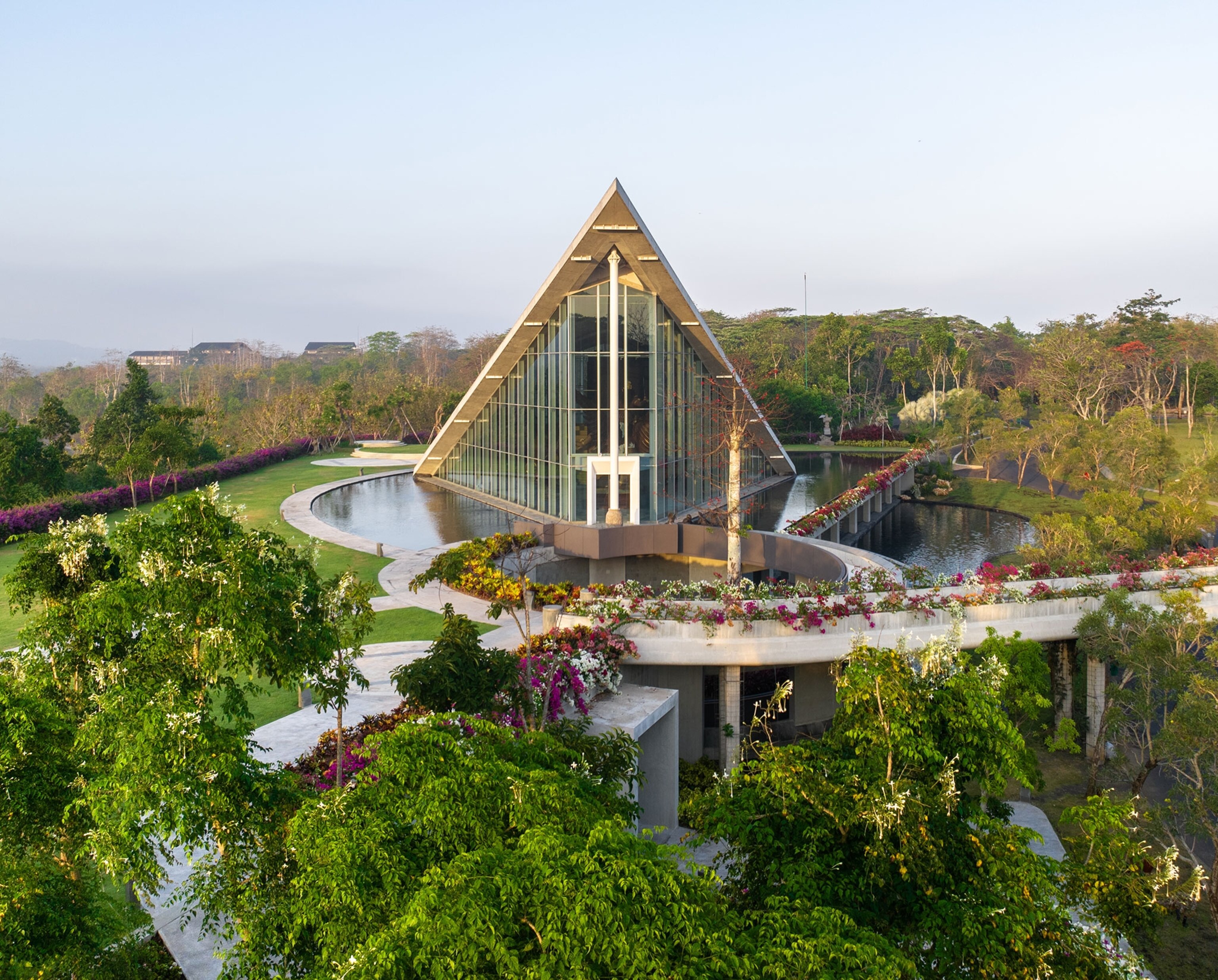 A triagular building, with two glass sides, is surround by several ponds and luscious green grass fields, tropical trees and plants.