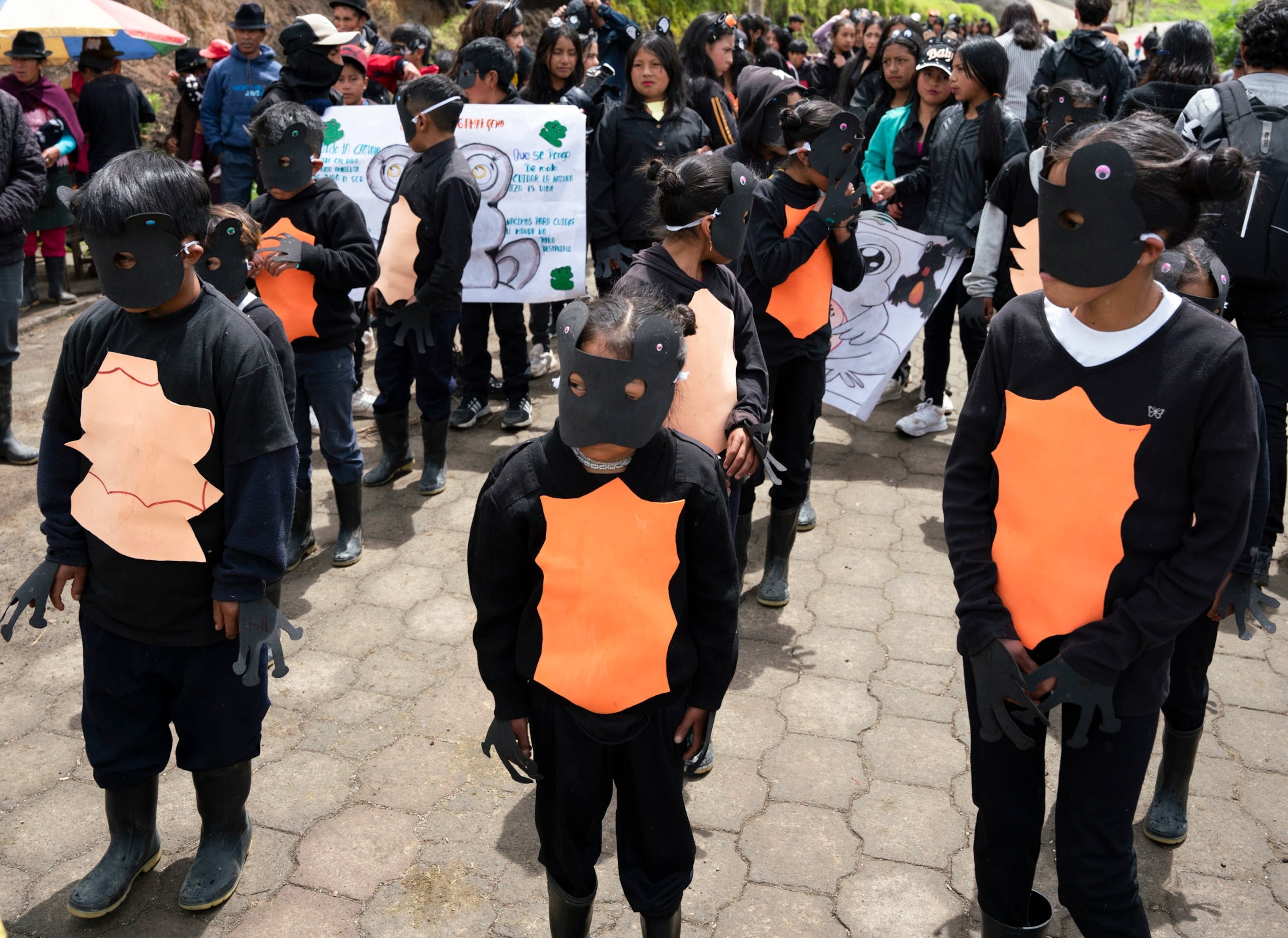 Three kids wearing black with orange on their stomachs are dressed as a toad, with others in the background.