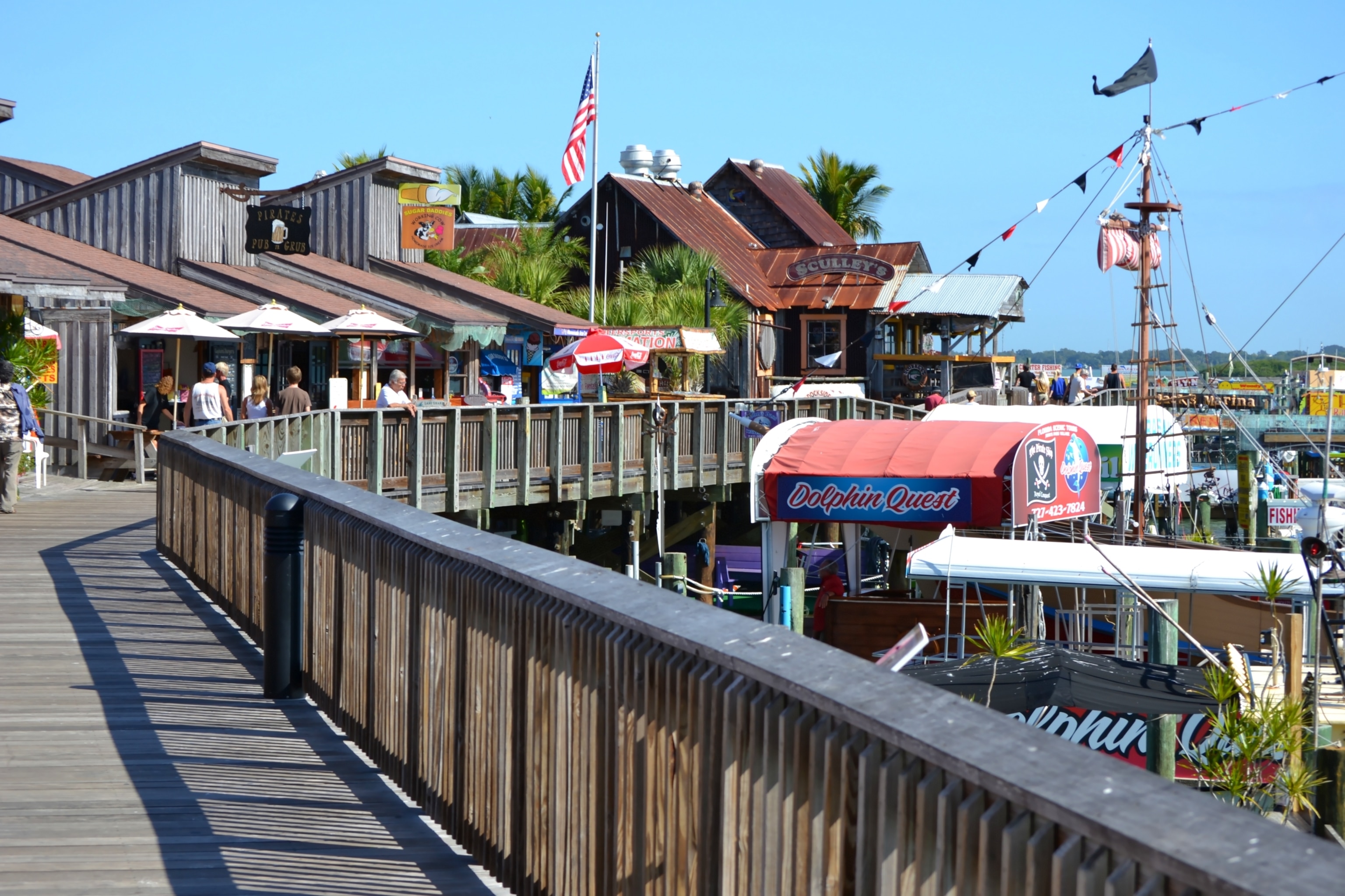 Boardwalk backed by wooden huts and shops with umbrellas and tables outside, while the boardwalk overlooks a marina lined with boats