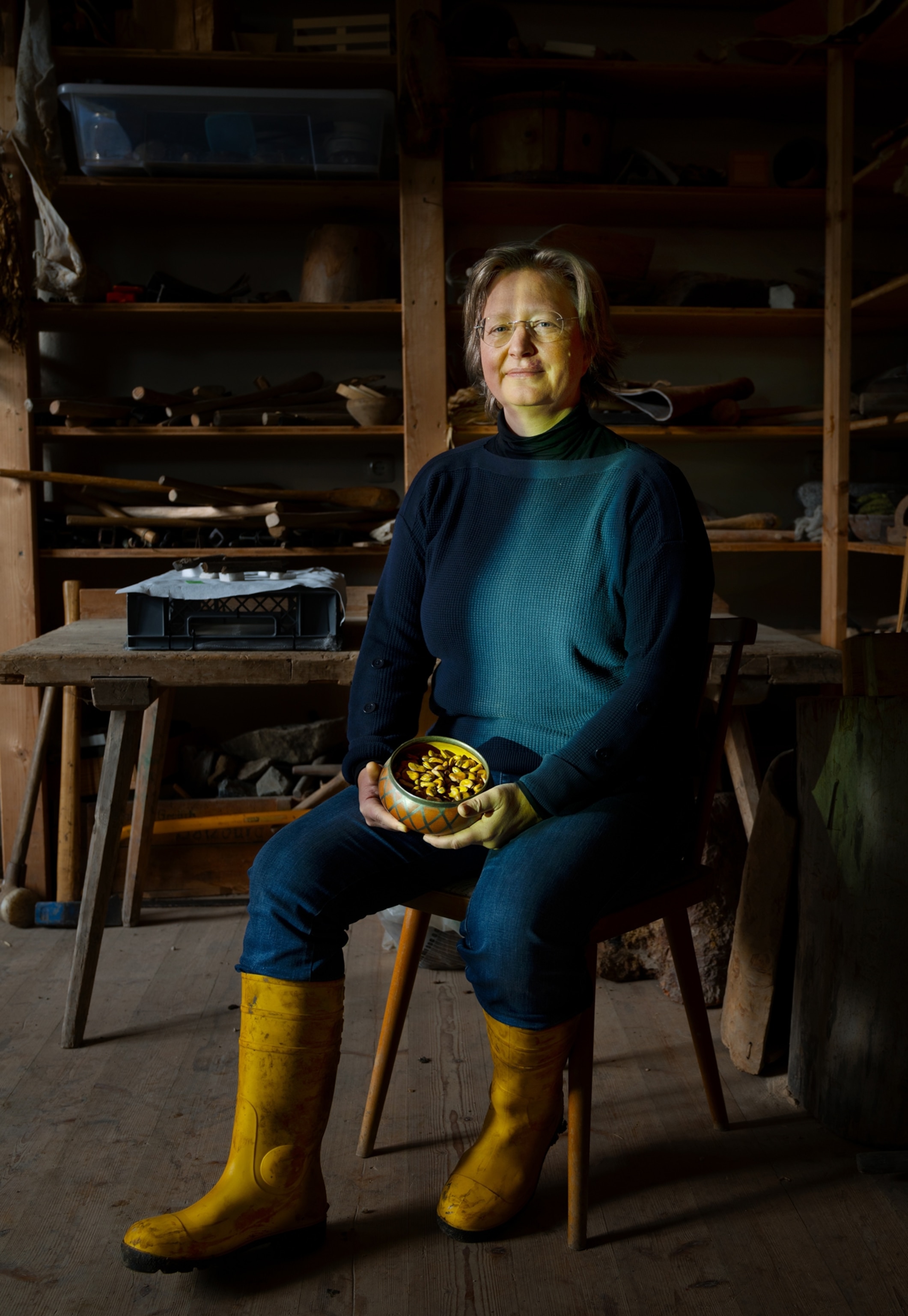 A woman sits in a chair in the middle of a room primarily made out of wood. She is wearing a blue sweat, blue pants, and yellow rain boots. In her hands she is holding a medium sized orange bowl filled with fava beans. Behind her is a wooden table and shelves along the wall with various items.