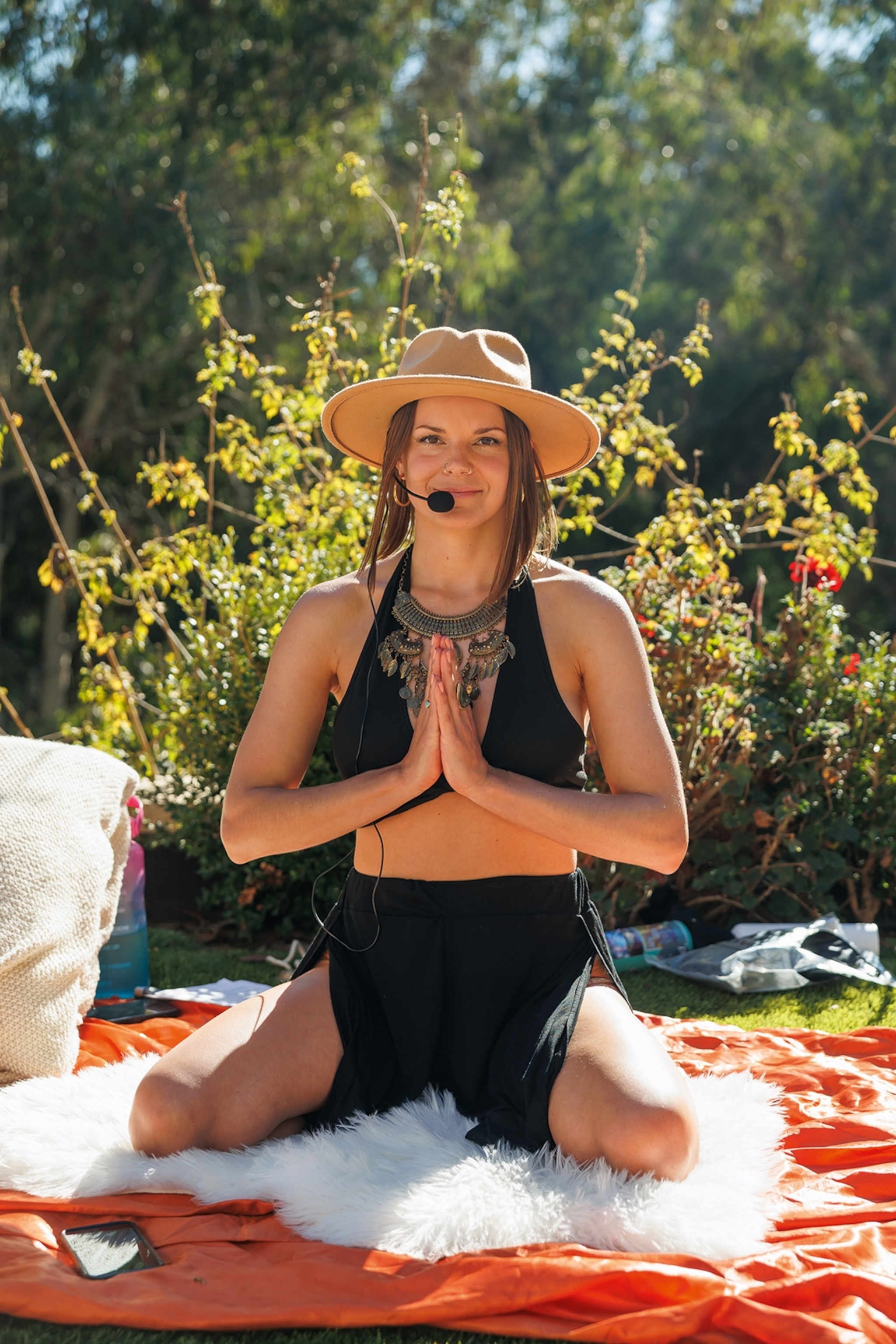 A young woman wearing yoga attire, a fedora hat and microphone, sitting outside with her legs crossed and her palms together in front of her chest.