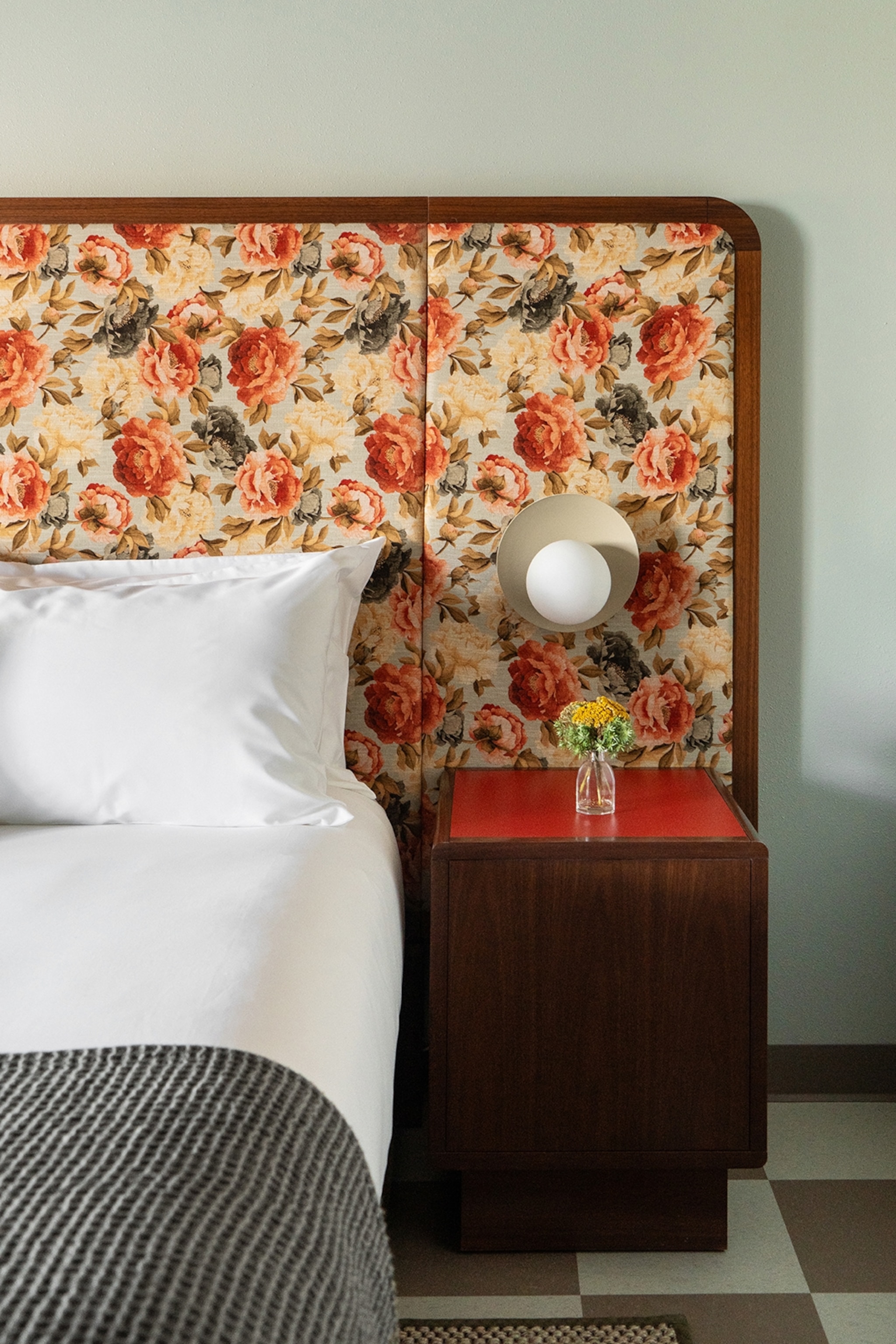 A bedside-table-view of a hotel bed with a floral-patterned headboard.