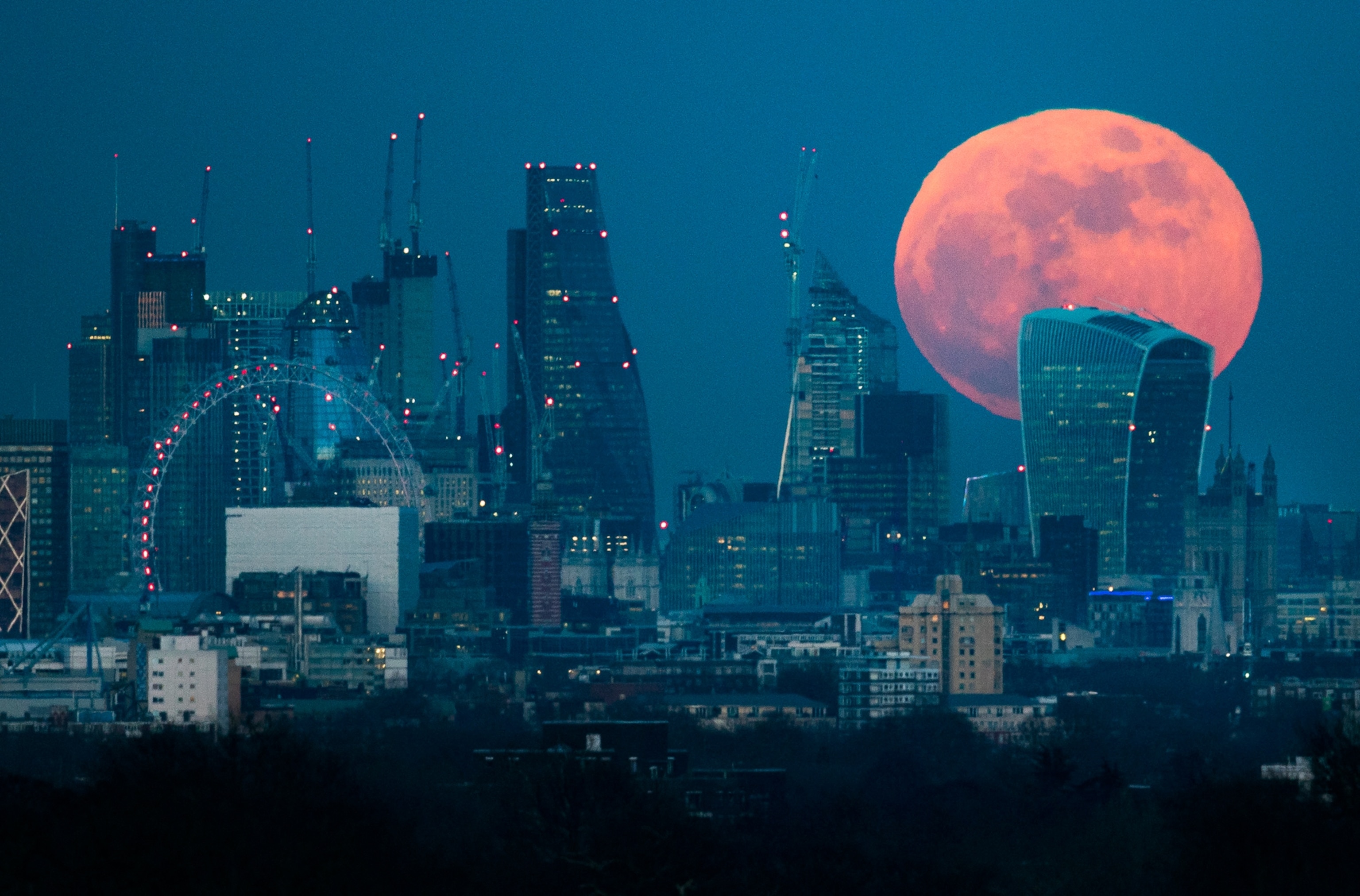A large, orange full moon rises behind London's skyline, featuring modern skyscrapers and cranes.
