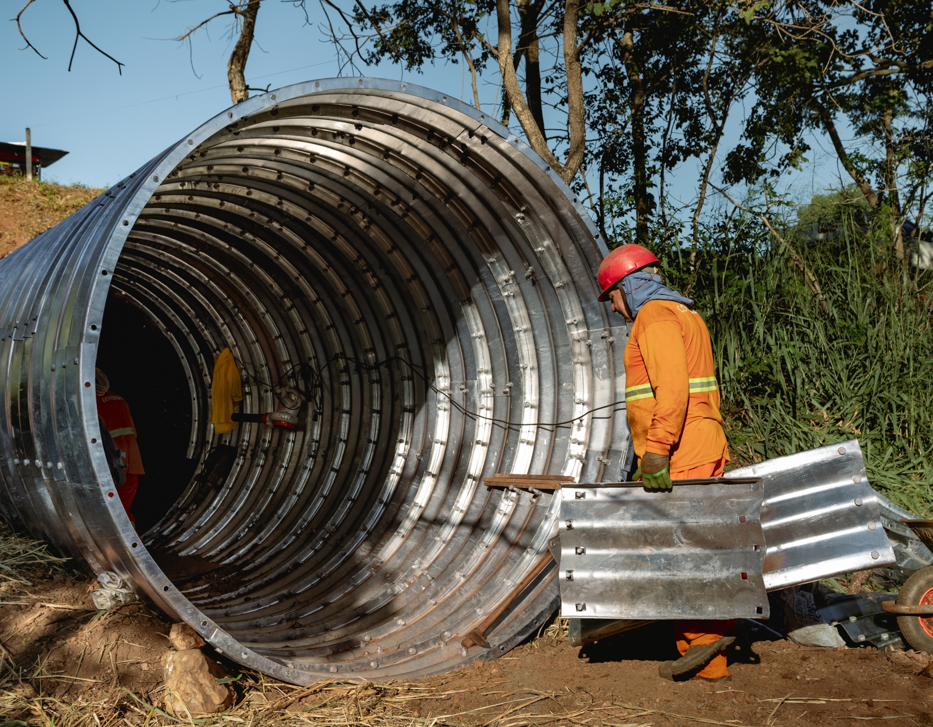 Workers wearing hard hands and orang jump suits walk into a large metal tunnel with additional rectangular tunnel piecing. The tunnel sits on dirt and goes under a dirt hill with a road above it. There is grass and trees spreading to the side of the tunnel's opening.