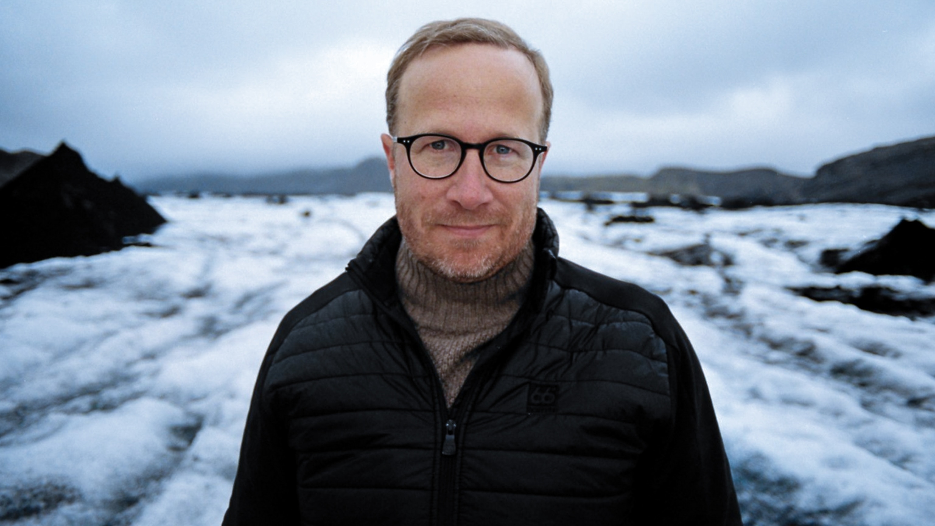 A man wearing glasses stands for a portrait amid a snowy landscape.