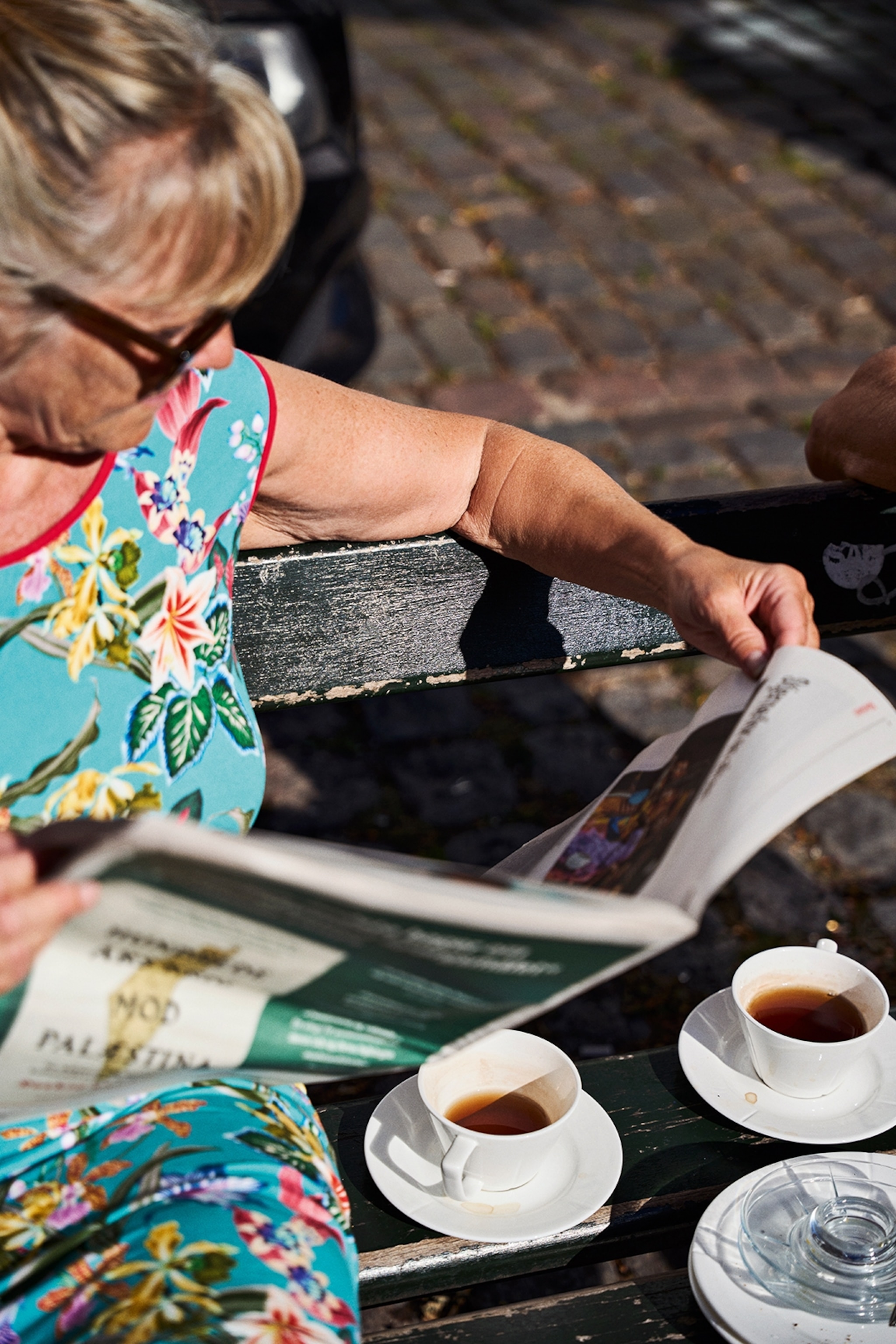A woman sat on a bench in the sun, reading the morning paper with a cup of coffee.
