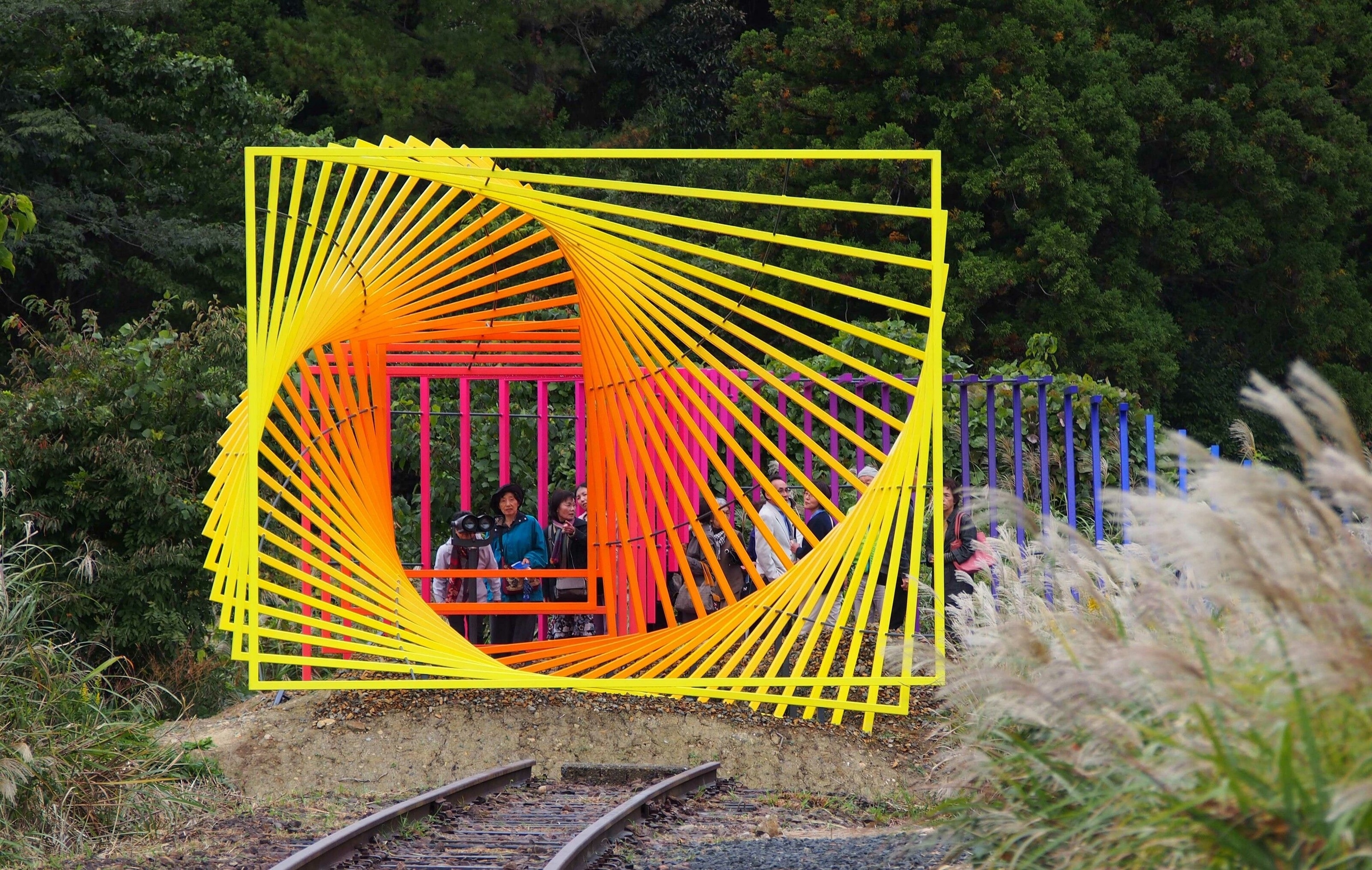 A piece of outdoor art. It is a metal spiral with a colour gradient that goes from yellow through orange, pink and purple all the way to blue.