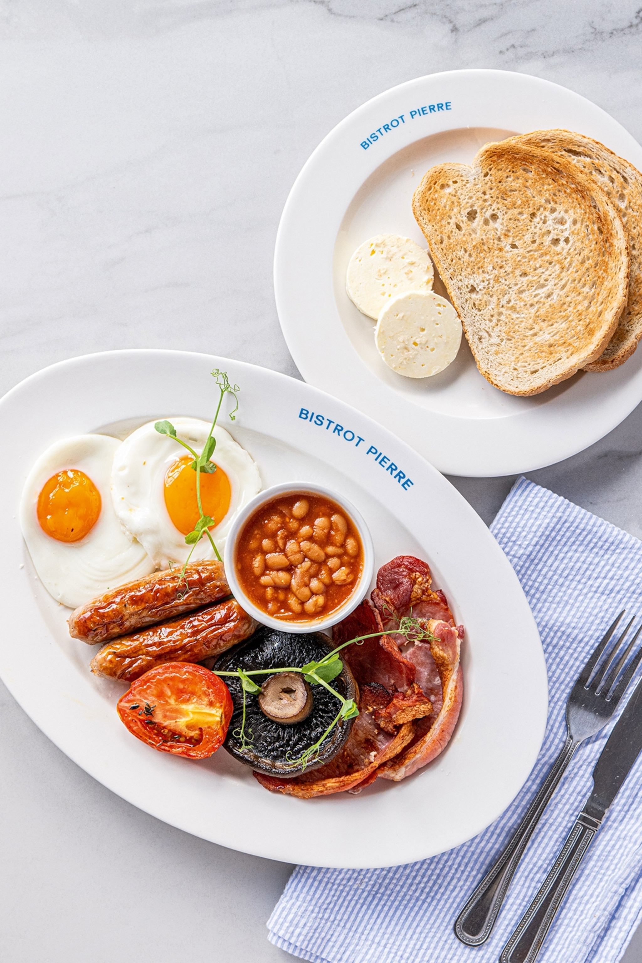 A plate of full English breakfast with fried egg, baked beans, sausages and two slices of bread on the side.