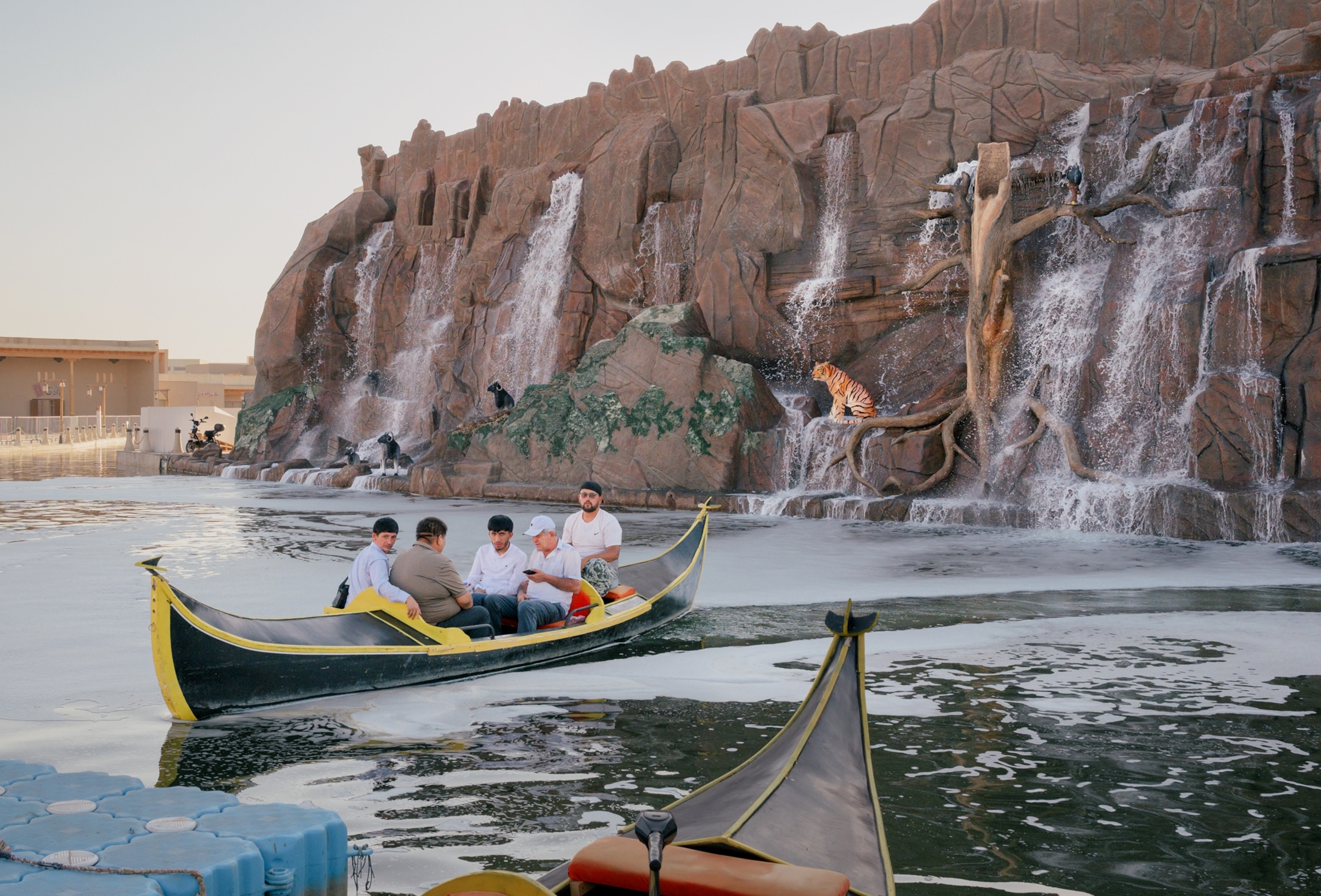 Two canoes sit in an artificial lake. A large, fake rock wall with waterfalls sits behind the lake in the background with a fake tiger sculpture sitting on the wall.