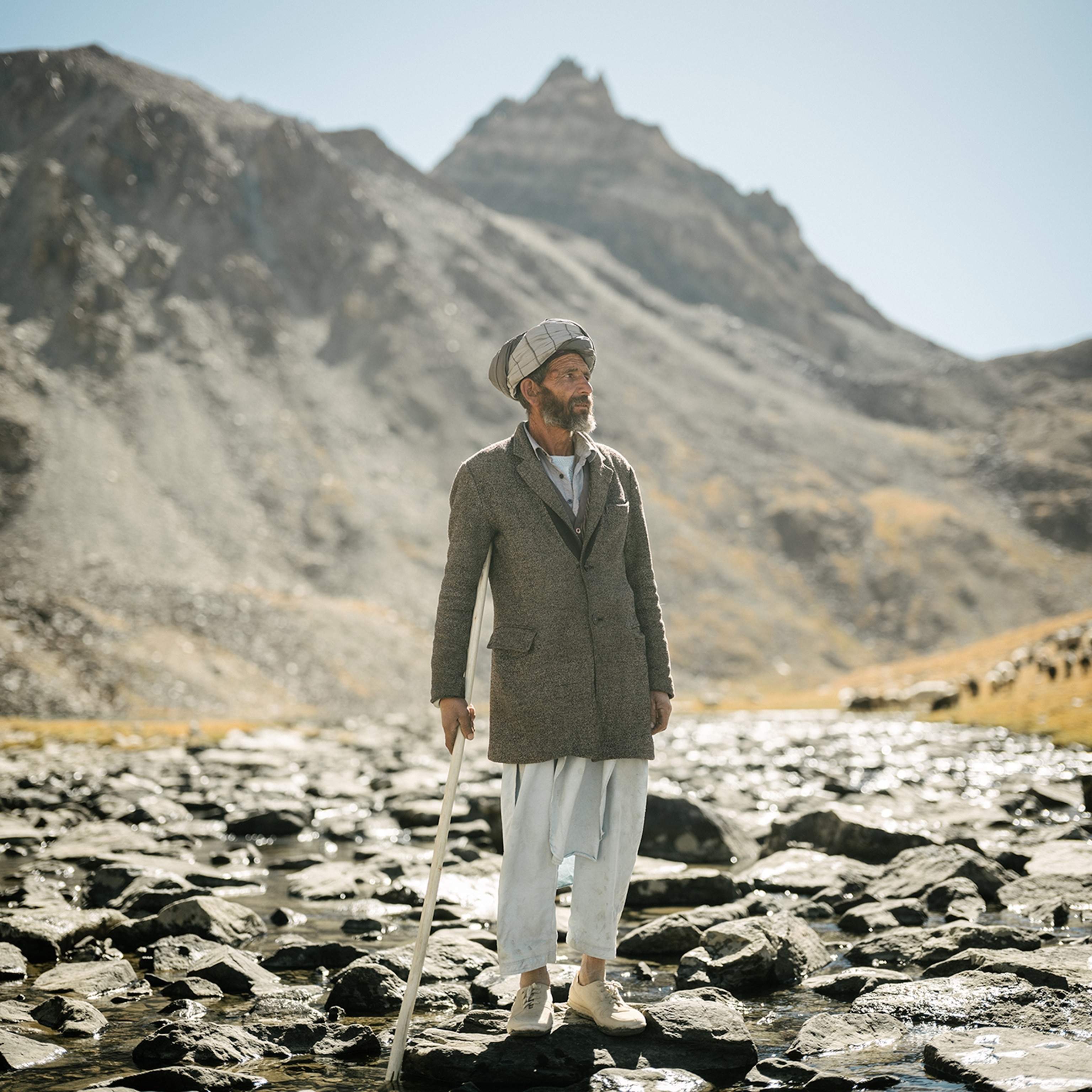 A man stands on rocks in a river and carries a pole. Behind him are mountains and livestock