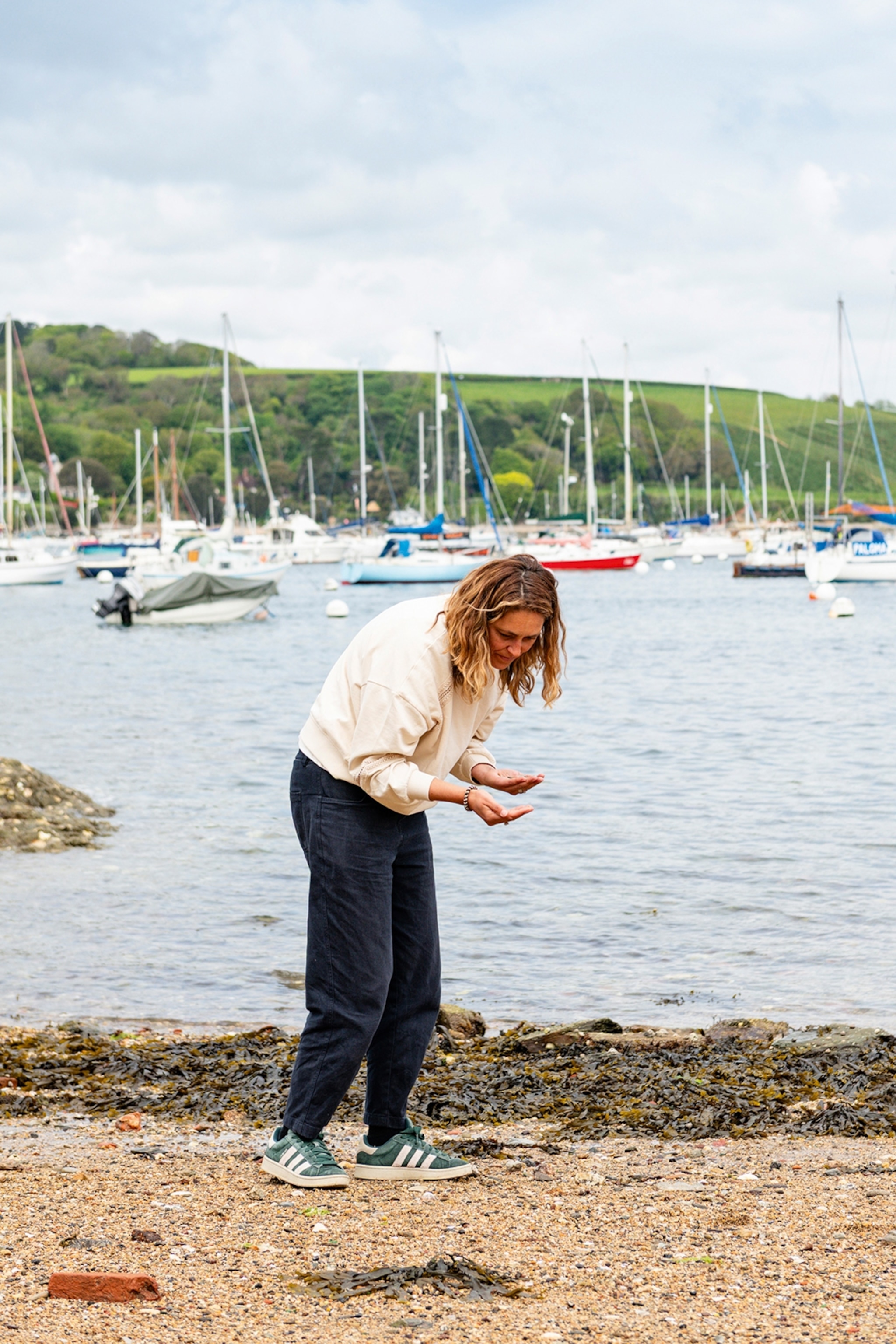 A woman bowing forward at a pebbled beach, looking at the palm of her hand with boats in the background.