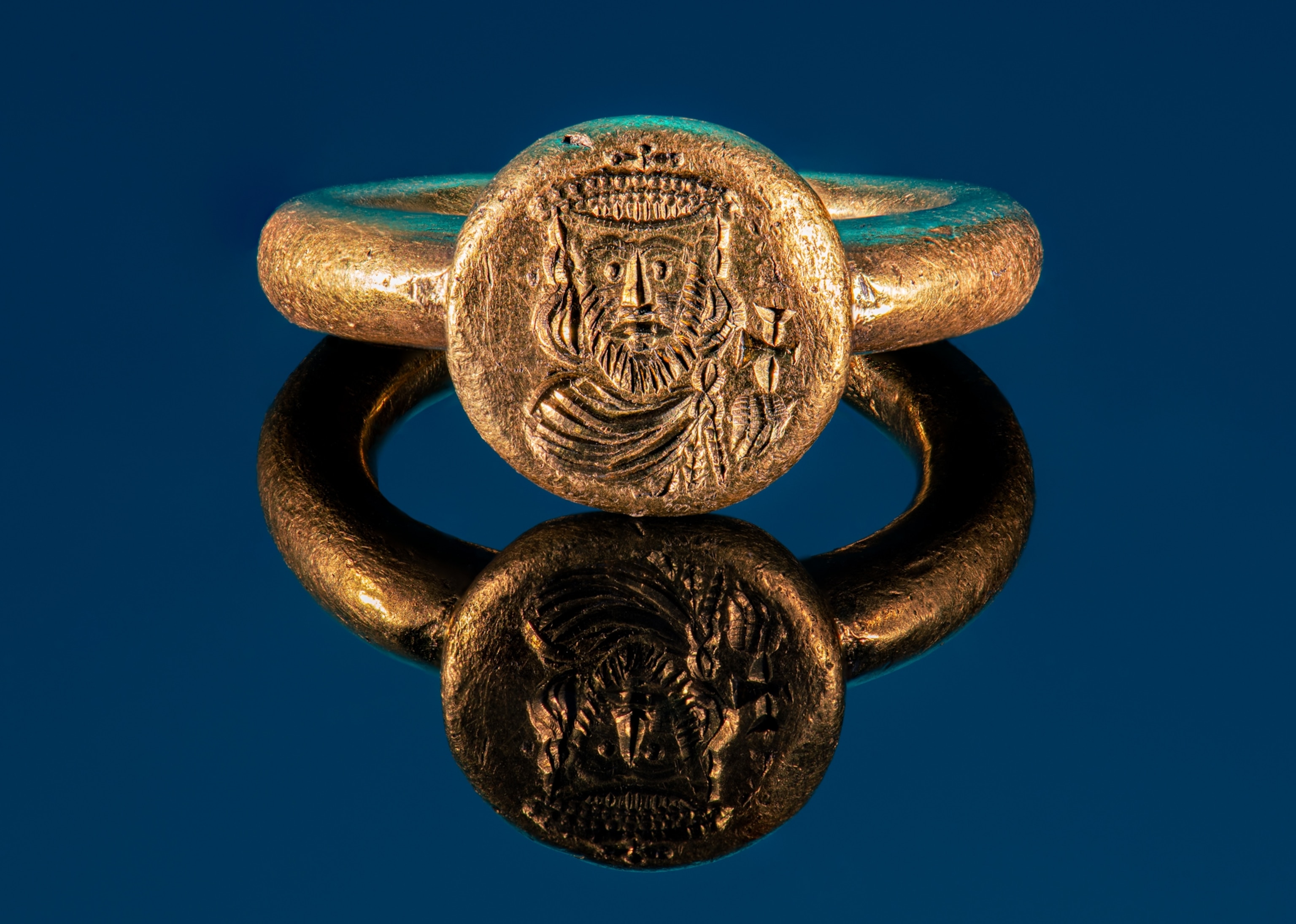 A gold ring with a round top. Stamped into the round piece is an illustration of a man with a beard wearing a crown and robes.