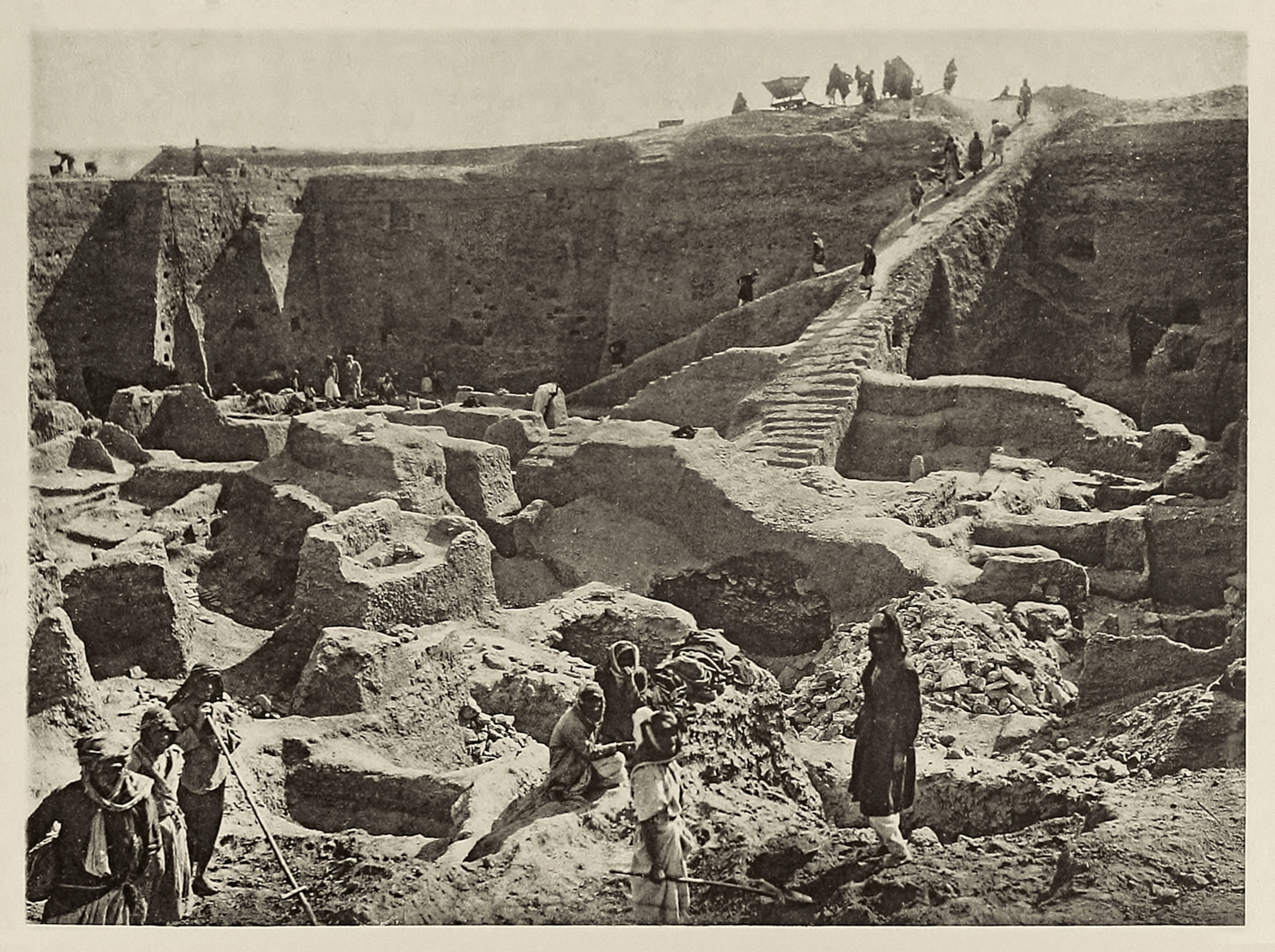 Archaeological excavation site with ancient ruins and stone structures. People are working, some walking up a carved staircase.