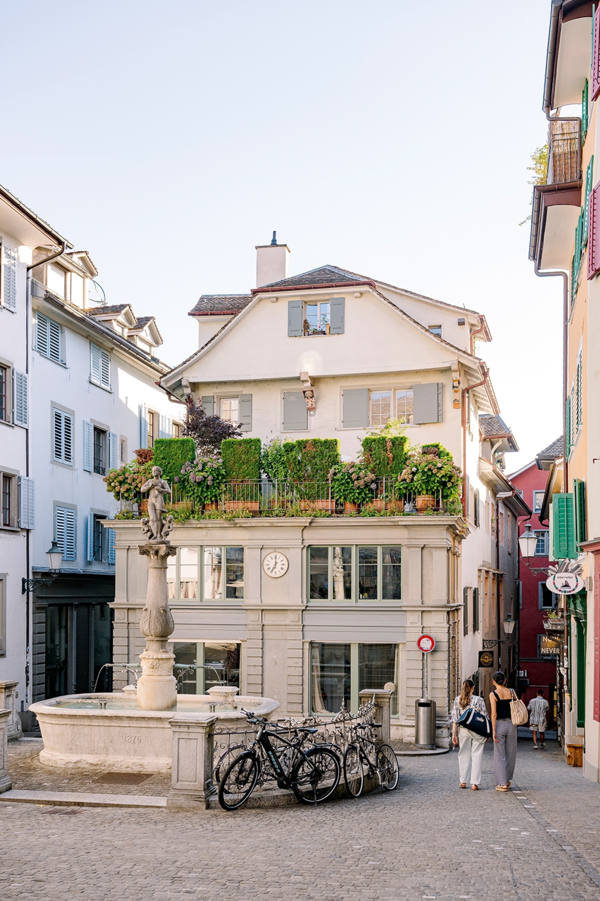 A charming plaza and small fountain in front of a cobble-stoned alleyway fork and luxurious, traditional house with wooden shutters.