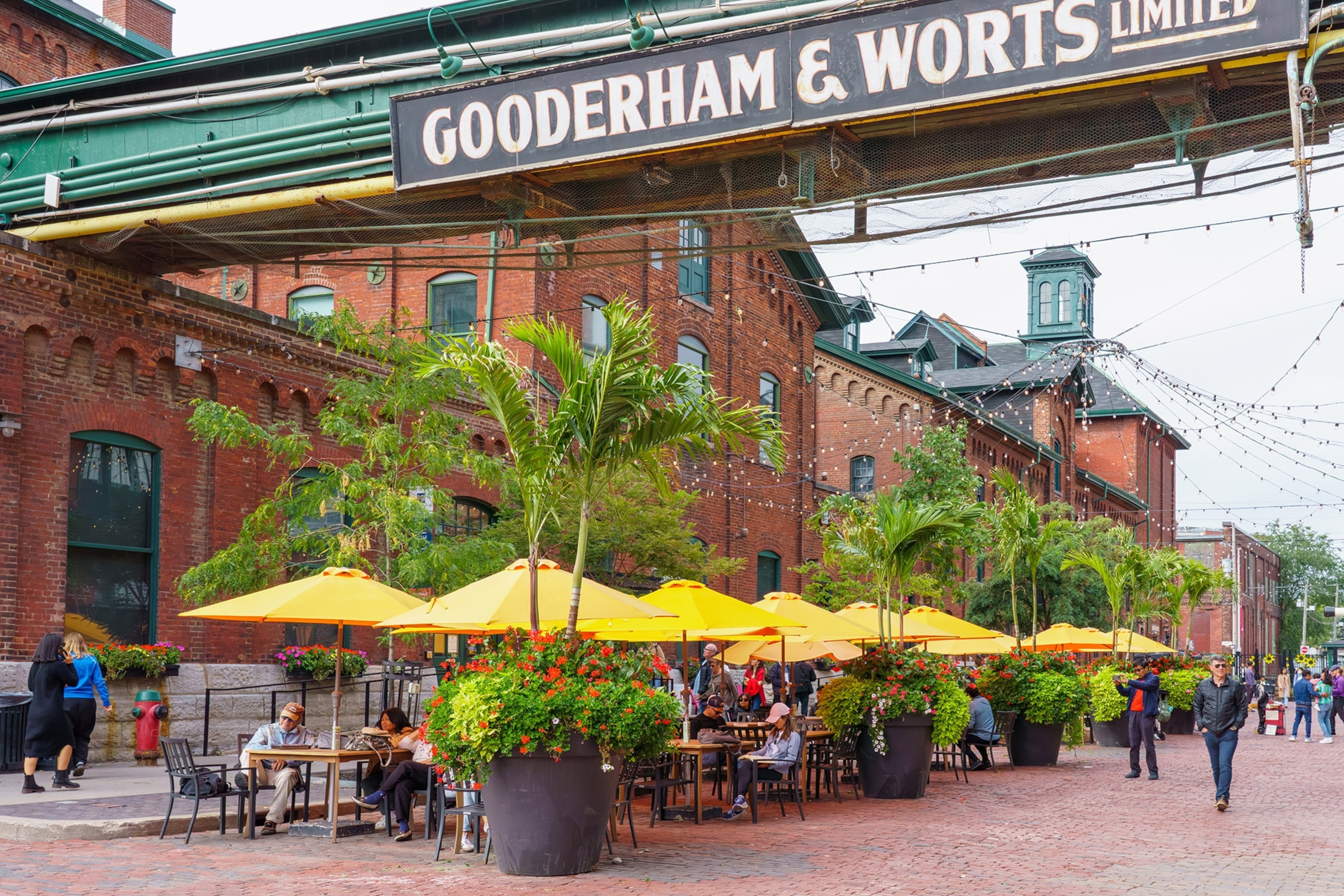 An industrial outdoor space with red brick buildings and red brick road under a steel beam reading "Gooderham & Worts", people sitting at an outdoor cafe under red umbrellas