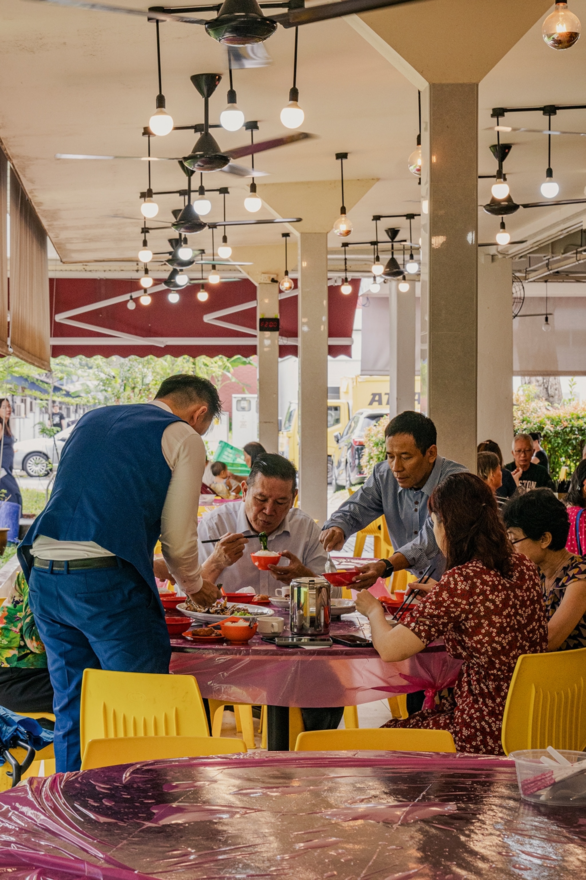 The dining patio of a traditional Malaysian restaurant with a plastic table cover and a family sharing multiple plates.