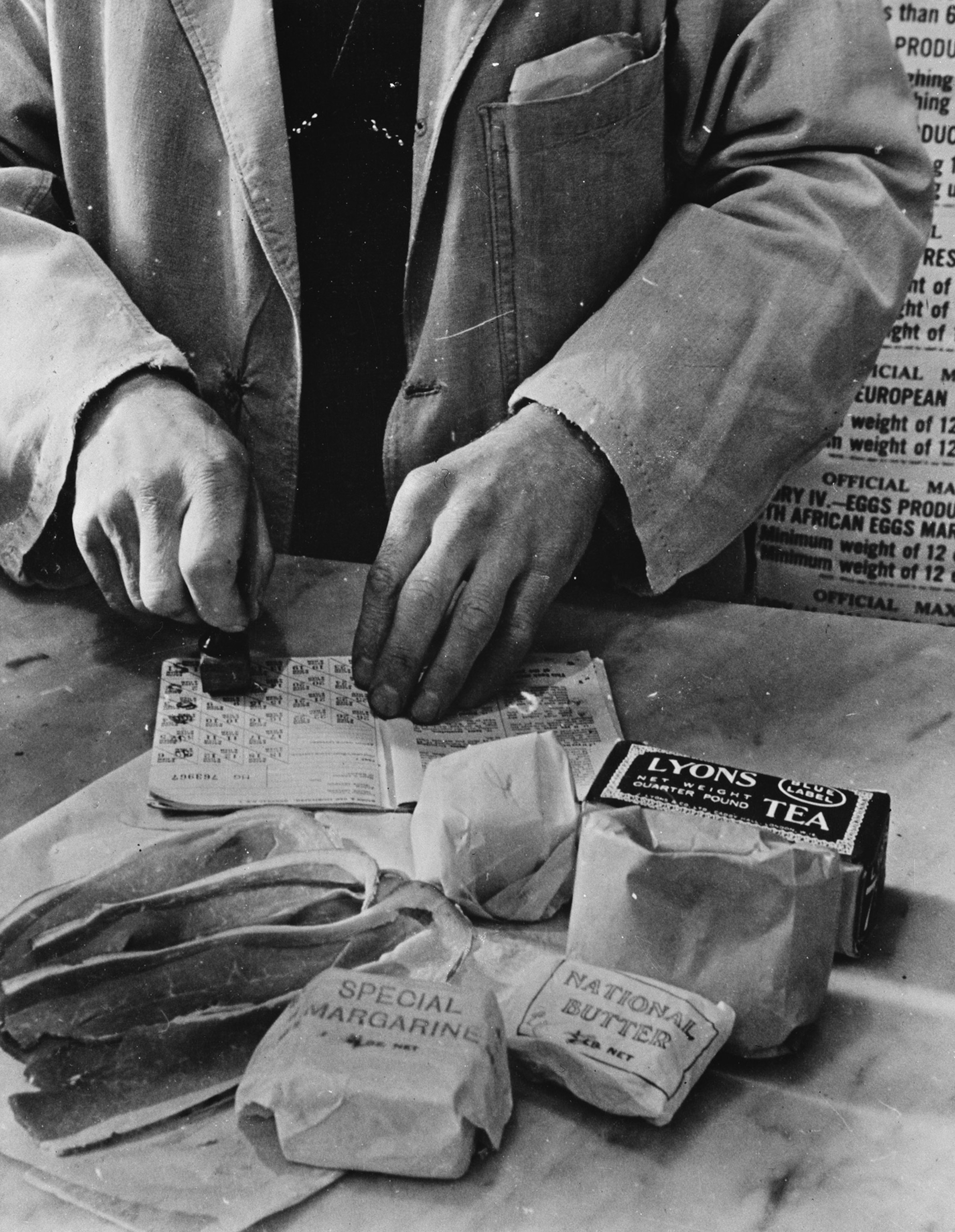 Black and white, an assortment of food items in foreground including packet labled "Special Margarine," a packet labeled "Lyons Tea," slices of bacon. In the background, an individual shown up to the chest, the hands holding a stamp and a booklet of postage-stamp size food ration notes.