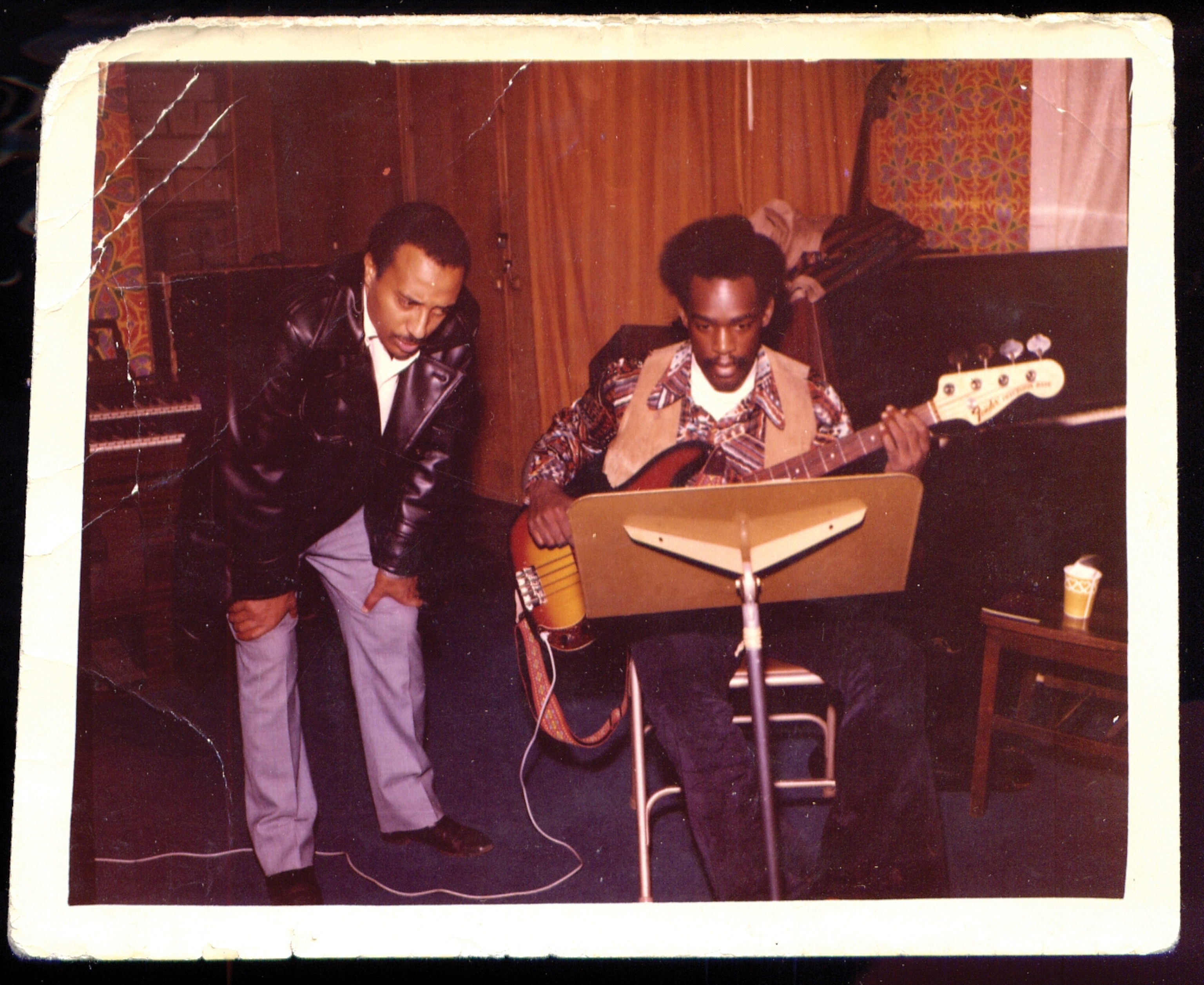 A man leans over looking at music while standing brancing himself another man sits in a chair playing an instrument.