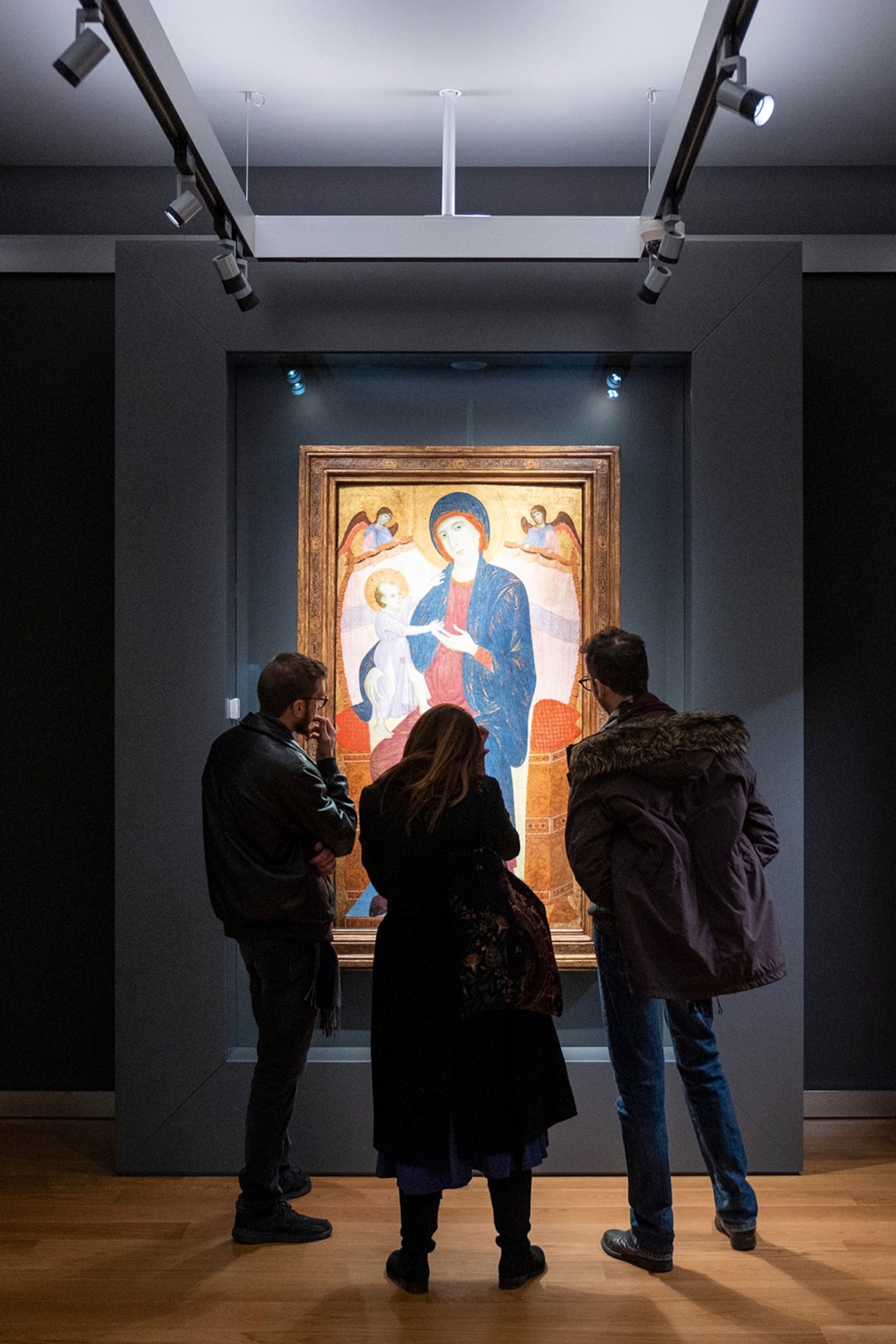 A religious portrait hung in a gallery and spotlit by two lamps as three visitors observe it.