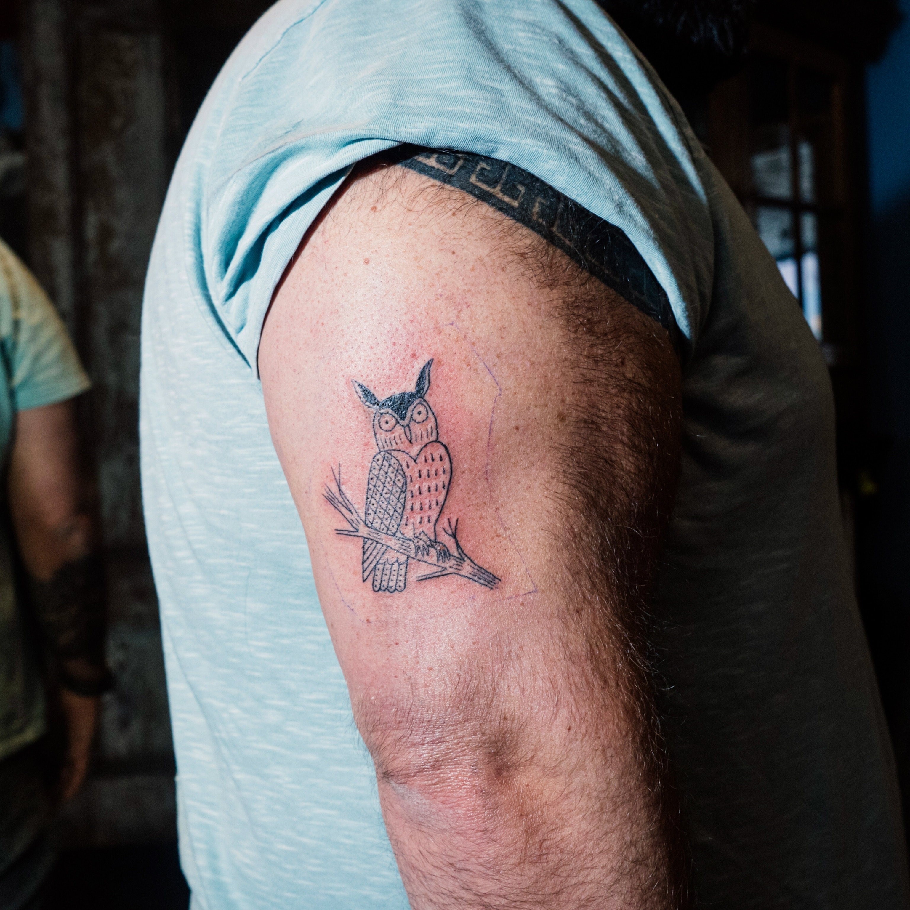 A person in a t-shirt displays a new owl tattoo on their upper arm. The owl is detailed and perched on a branch, while skin around the tattoo is irritated and red.