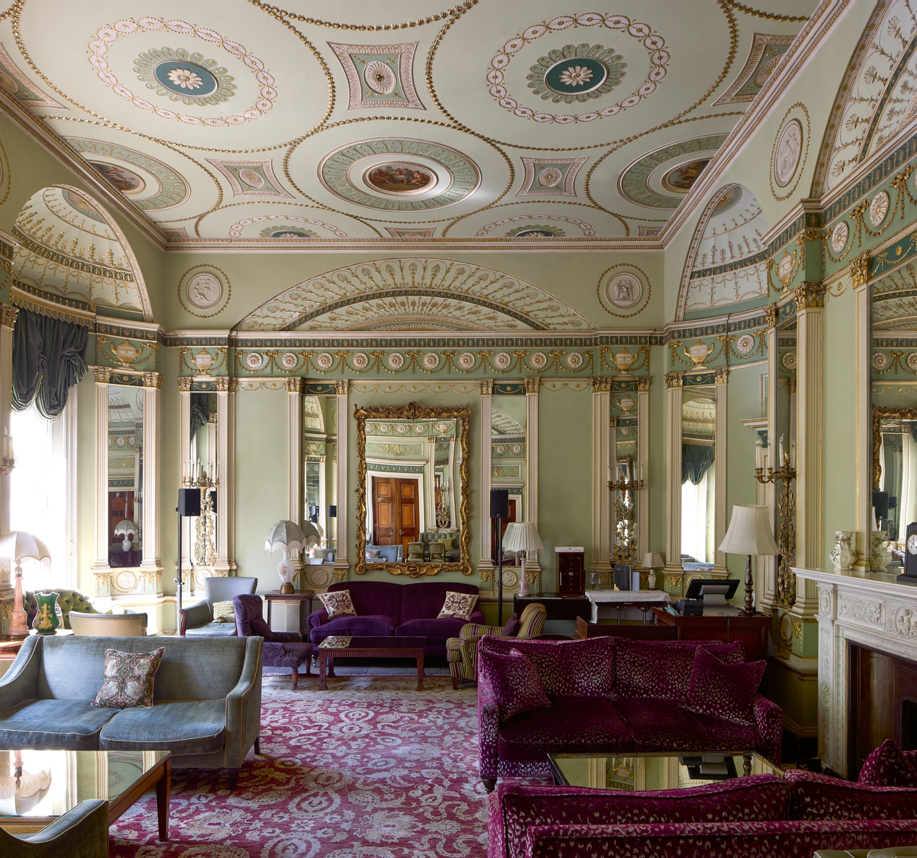 Elegant drawing room with ornately decorated ceiling and walls, mirrors, plush purple and grey sofas, and a patterned red carpet, exuding a regal ambiance.