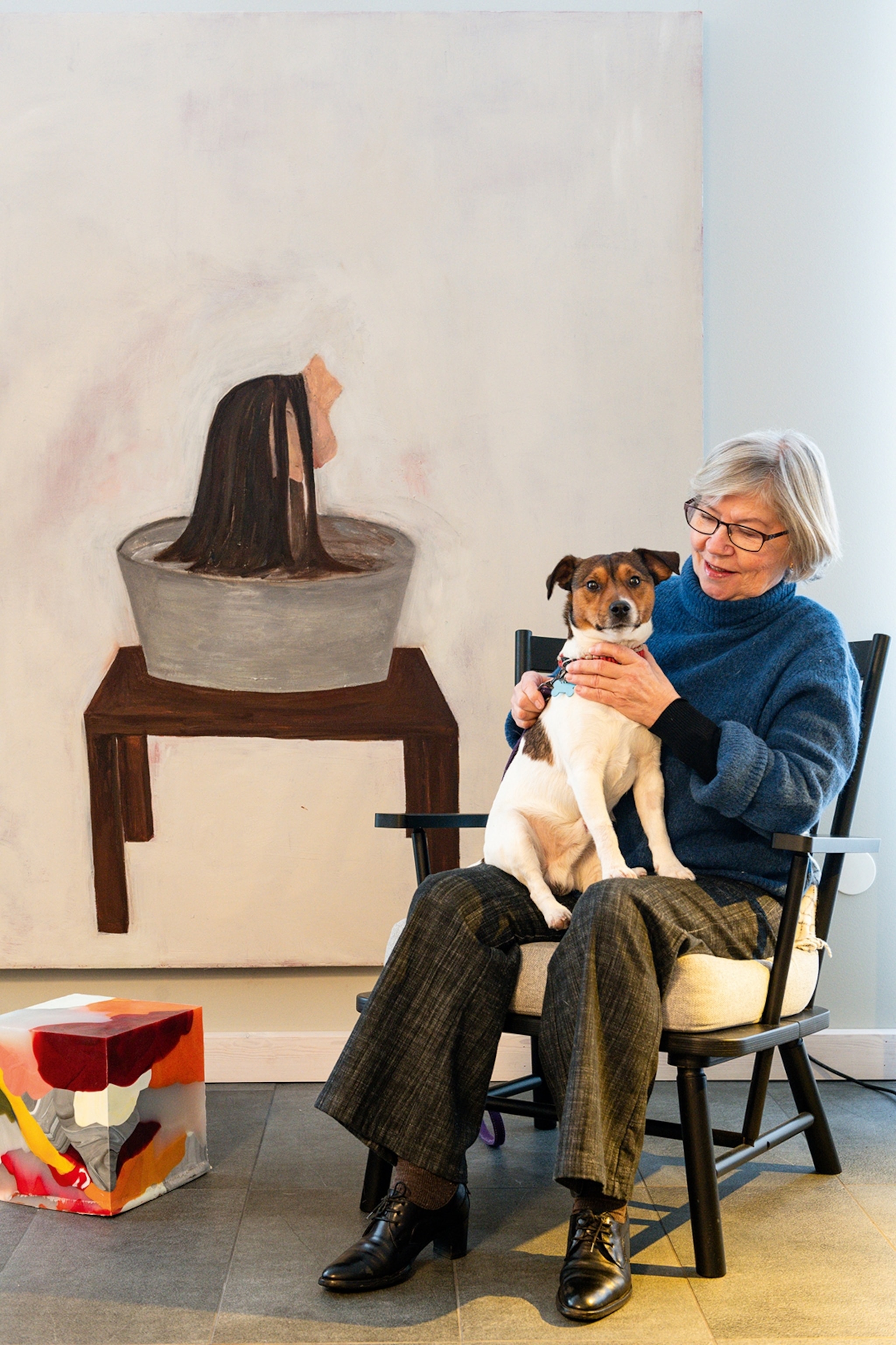 An elderly, smiling woman on a polstered wooden chair wit her terrier dog on her lap and art in the background.
