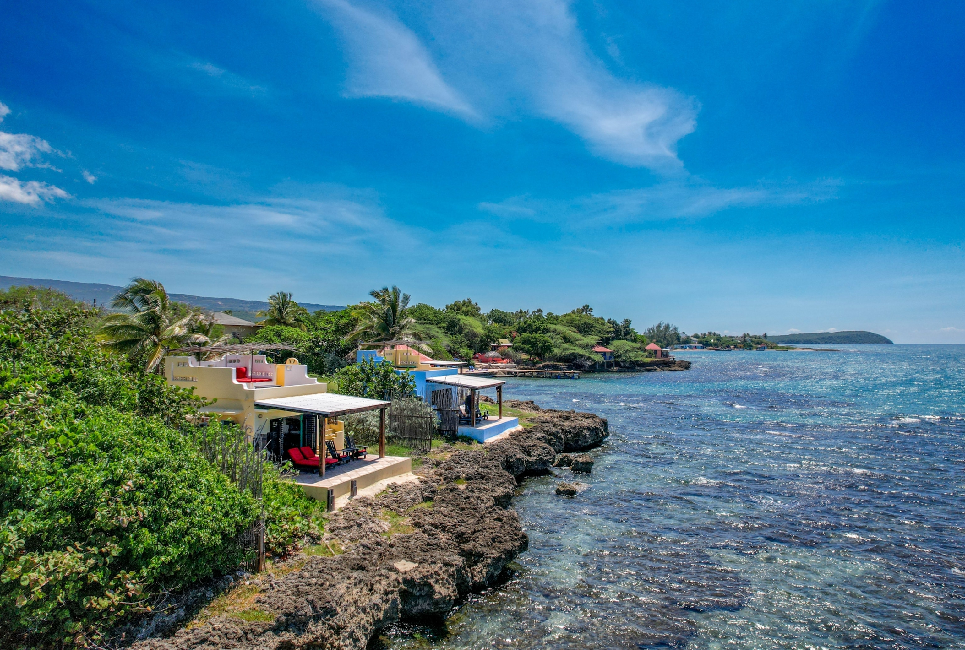 Colorful seaside villas sit along a rocky shoreline surrounded by tropical greenery, with clear blue ocean waters stretching into the distance.