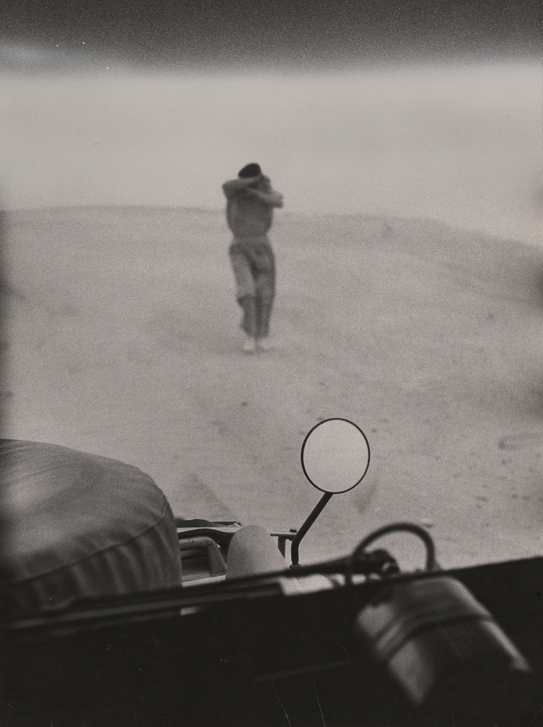 Lashed and blinded by a sandstorm, a man his way to a car whose mirror is in the foreground.