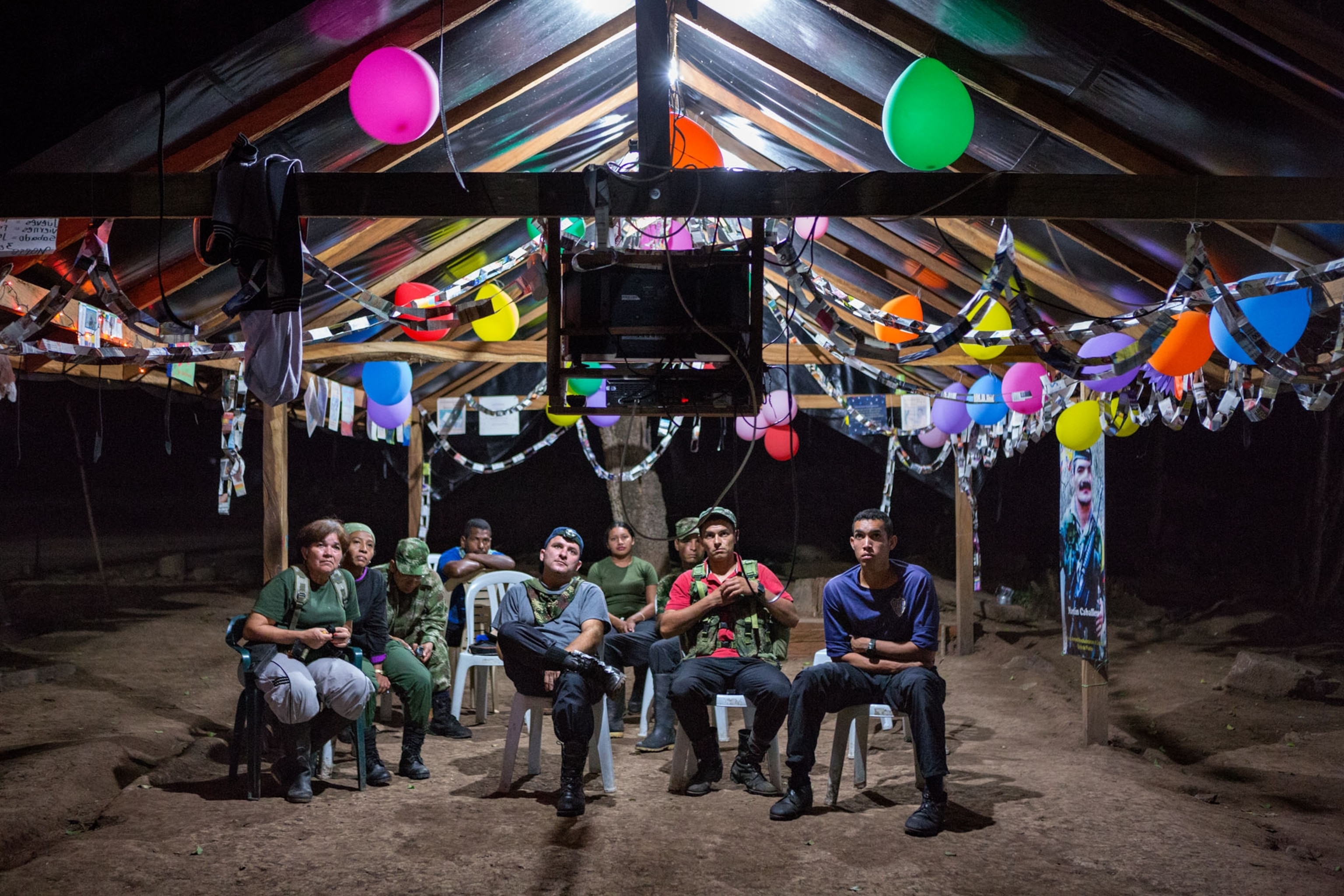 a group of men sitting outside watching tv at night as balloons hang above them