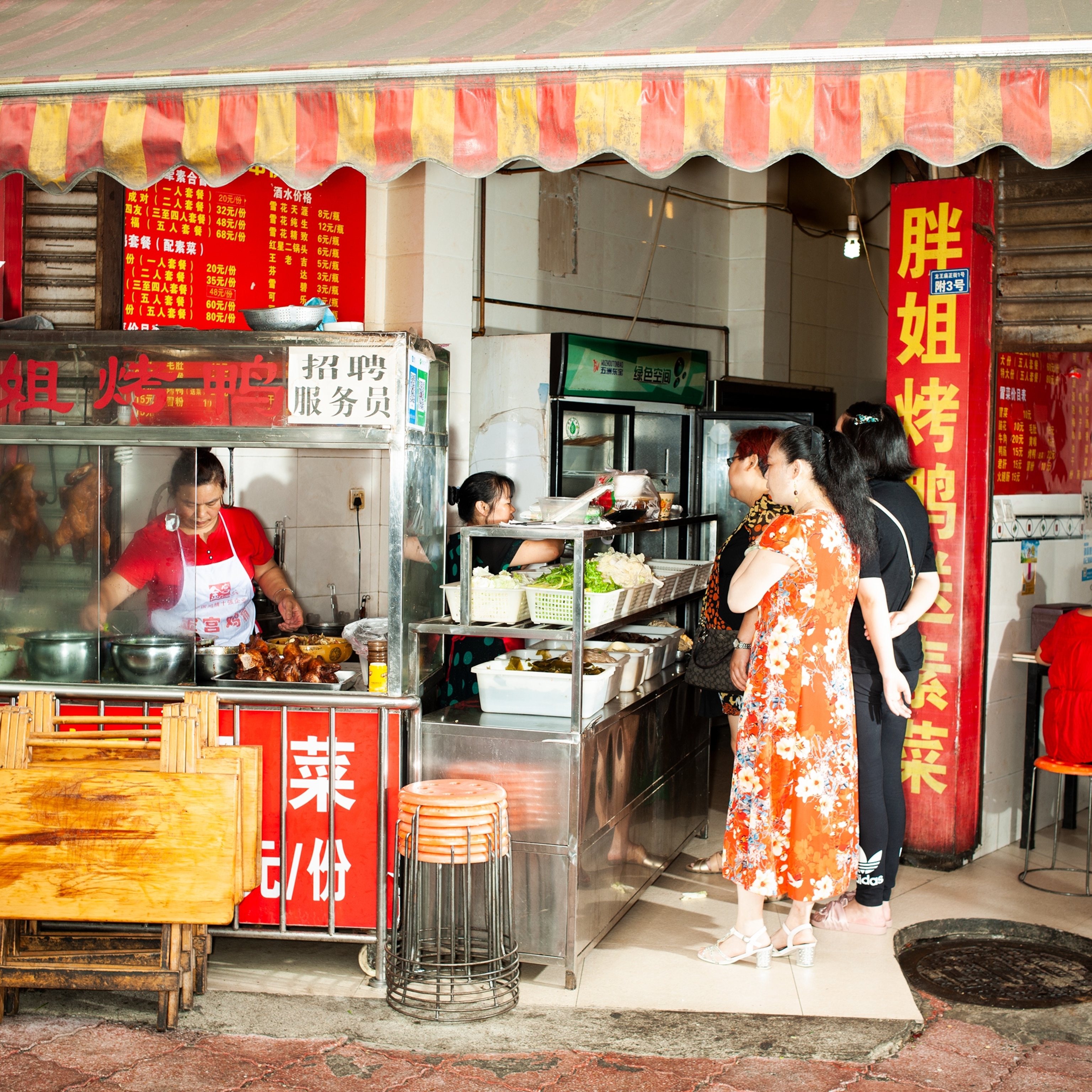 A street food stall serves customers at a small restaurant entrance, with trays of prepared dishes on display and bright red signs with Chinese characters hanging around the storefront.