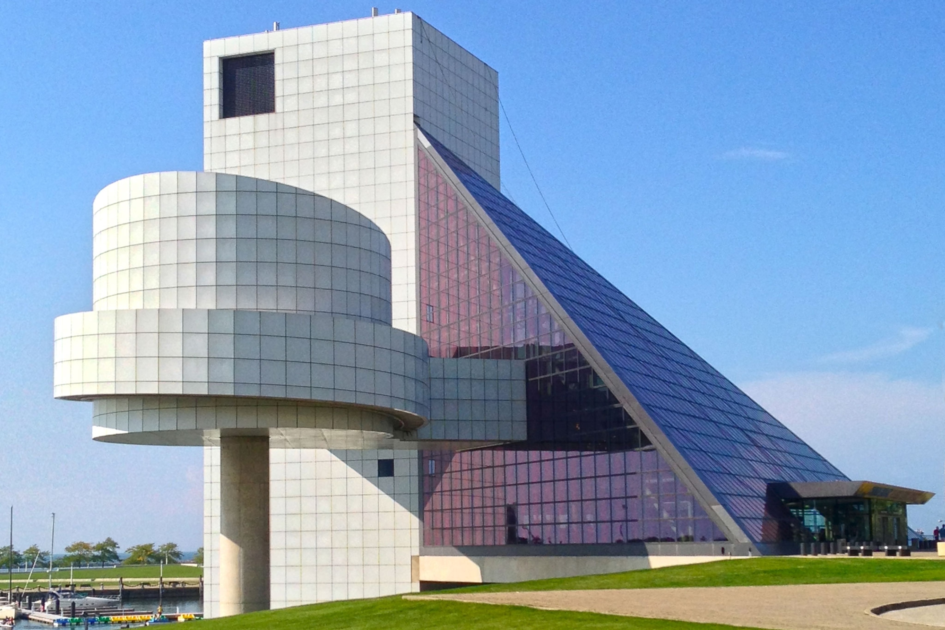 Alt text: Modern architectural building with a combination of geometric shapes, including a cylindrical structure in the front and a large glass pyramid in the back, against a clear blue sky. This is the Rock and Roll Hall of Fame in Cleveland, Ohio.