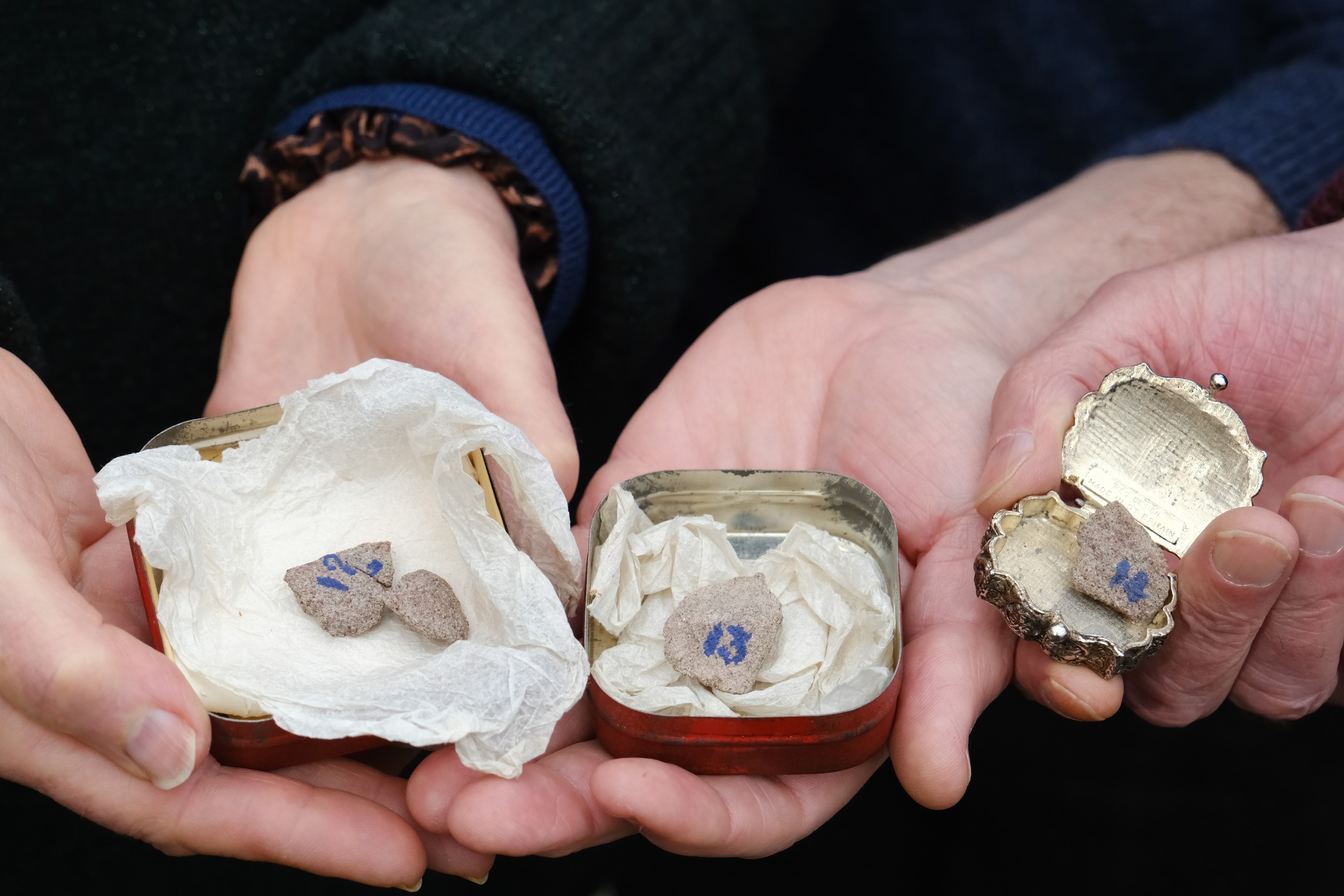 A group of hands hold fragments of stone in tins