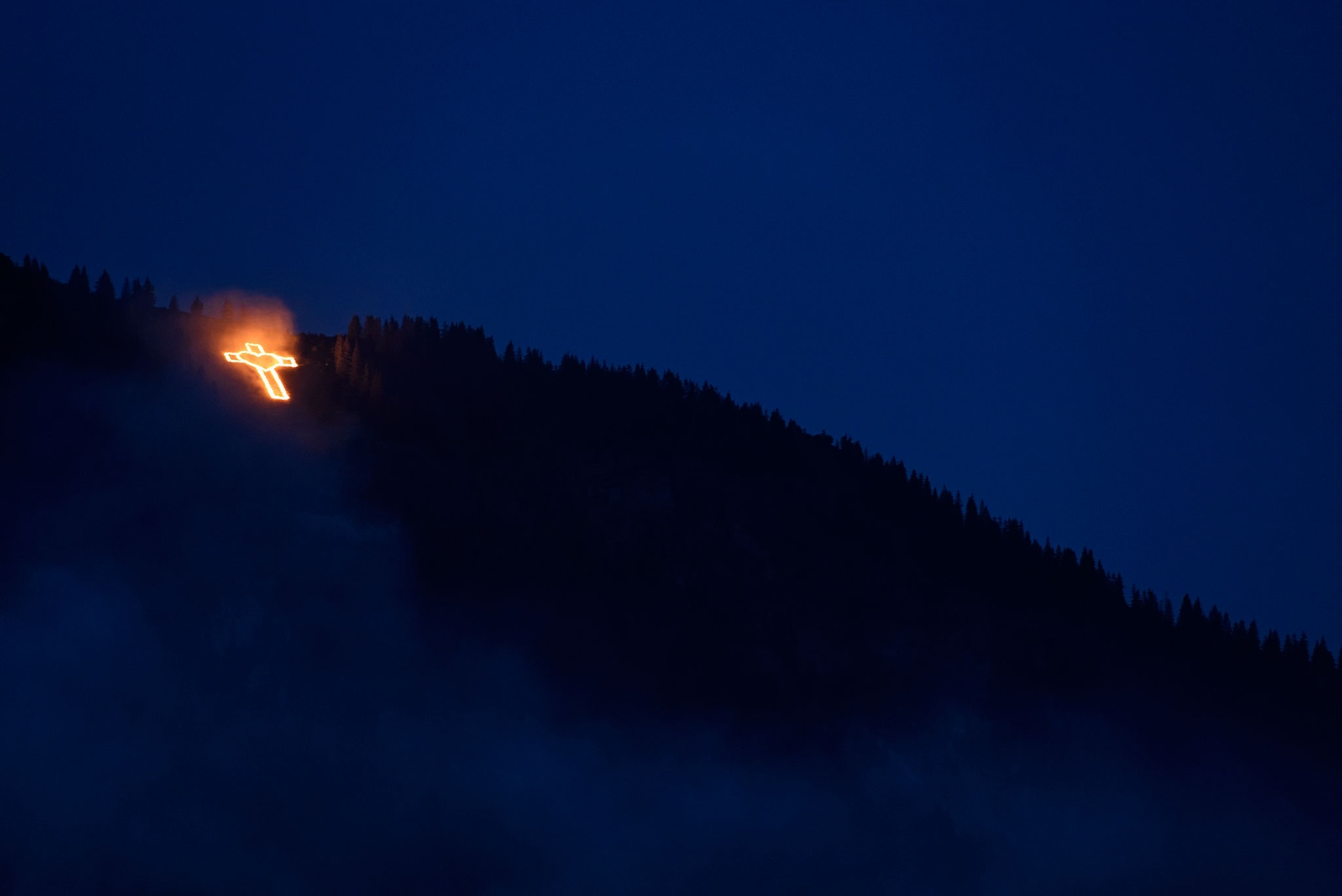 a cross lit up by fire on the side of a mountain in the Alps