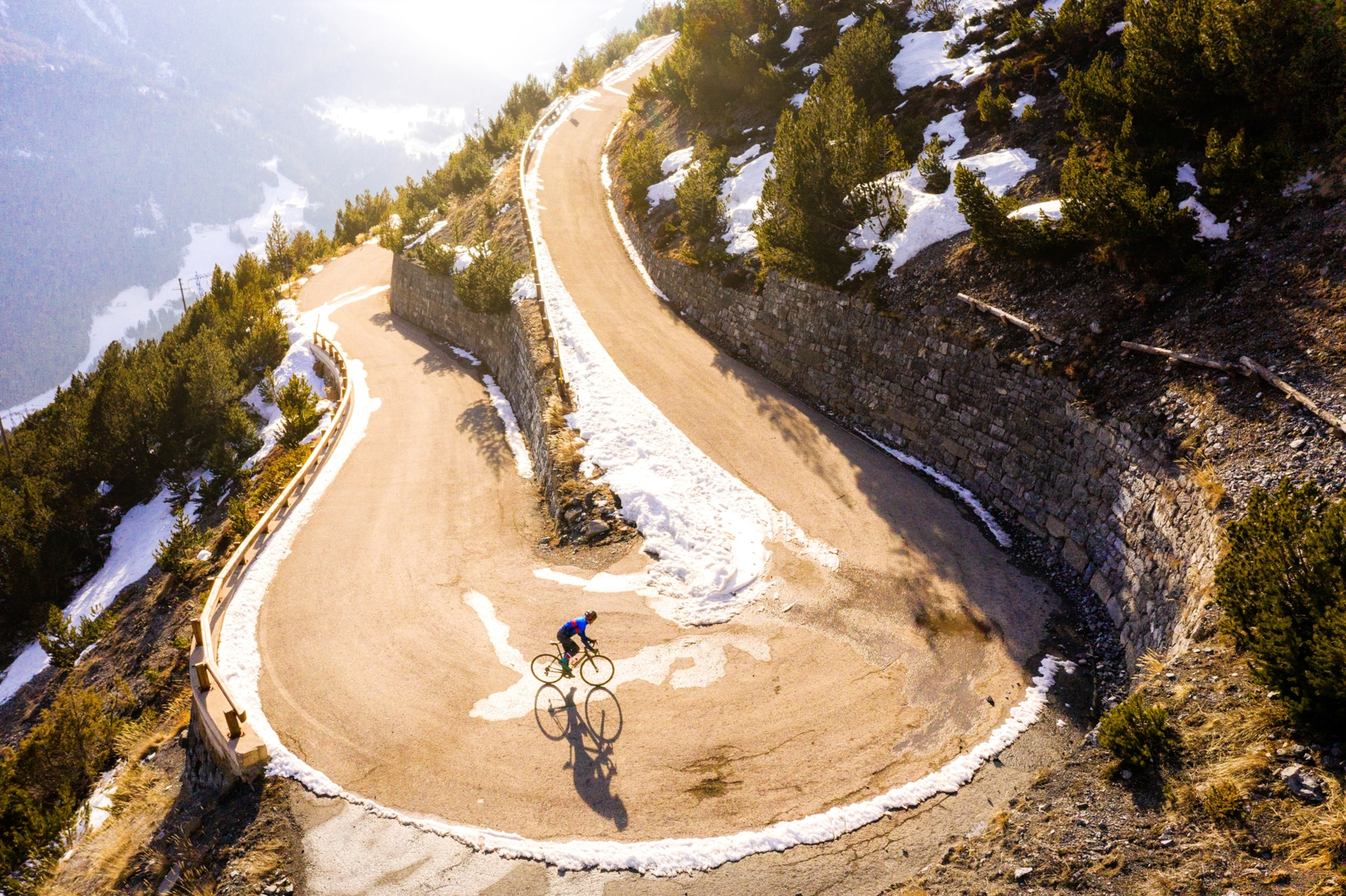 A cyclist in a blue jacket rides up a sunlit, snow-lined mountain road curve, surrounded by pine trees and rocky terrain