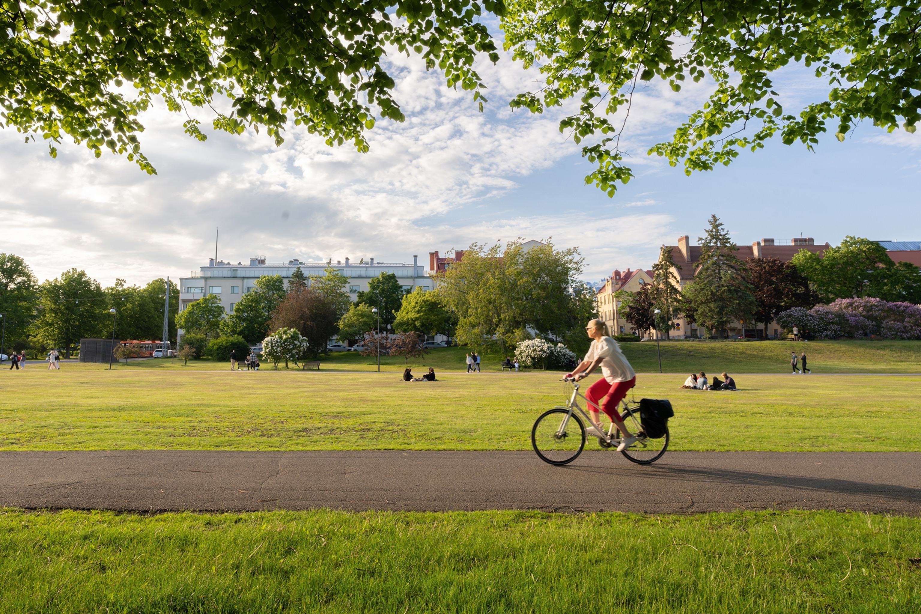 A wide, urban park landscape with a woman cycling past on a bike and more park-goers in the background.
