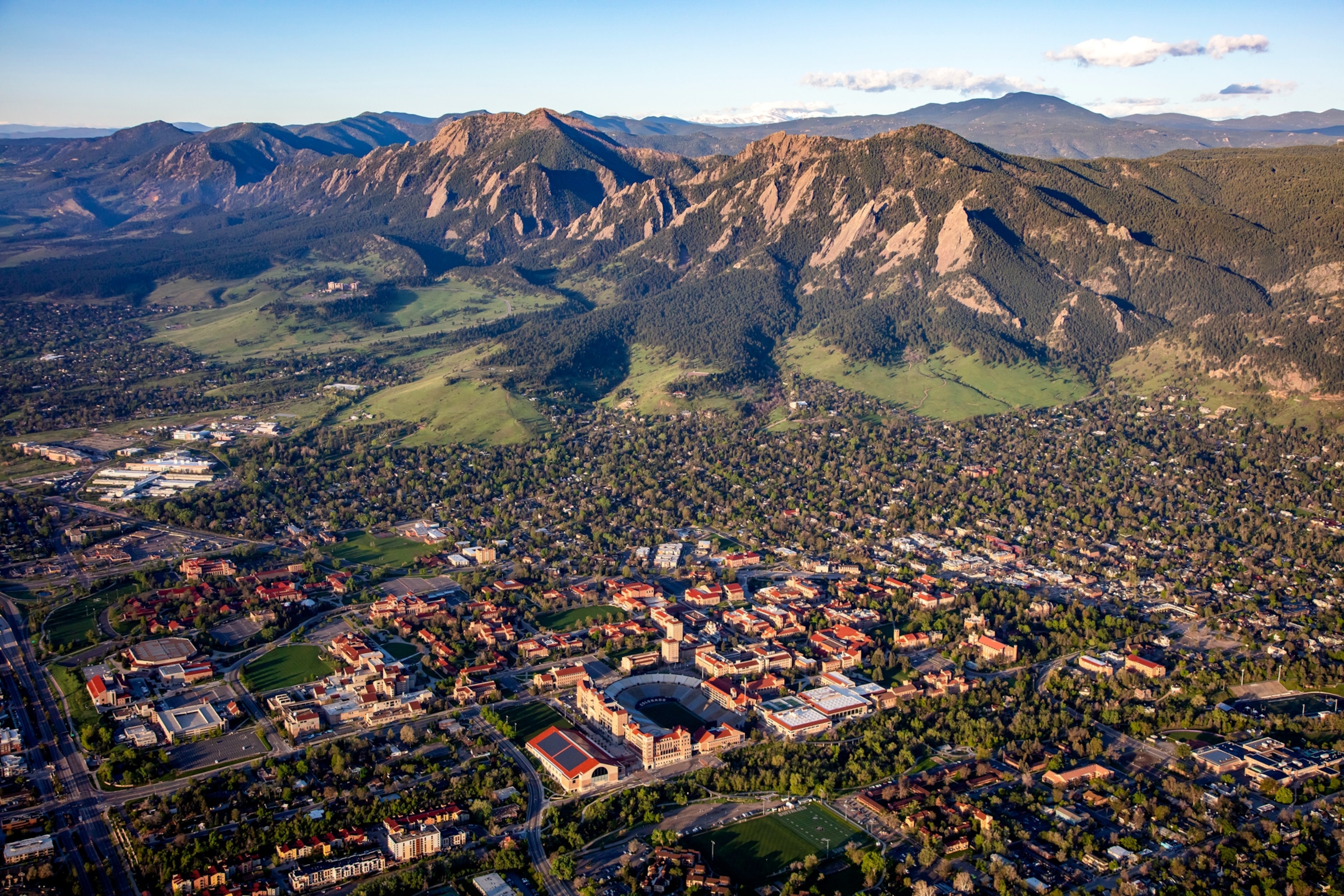 An aerial view of a city at the base of a rugged mountain range, with neighborhoods, roads, and buildings spread across the valley below.