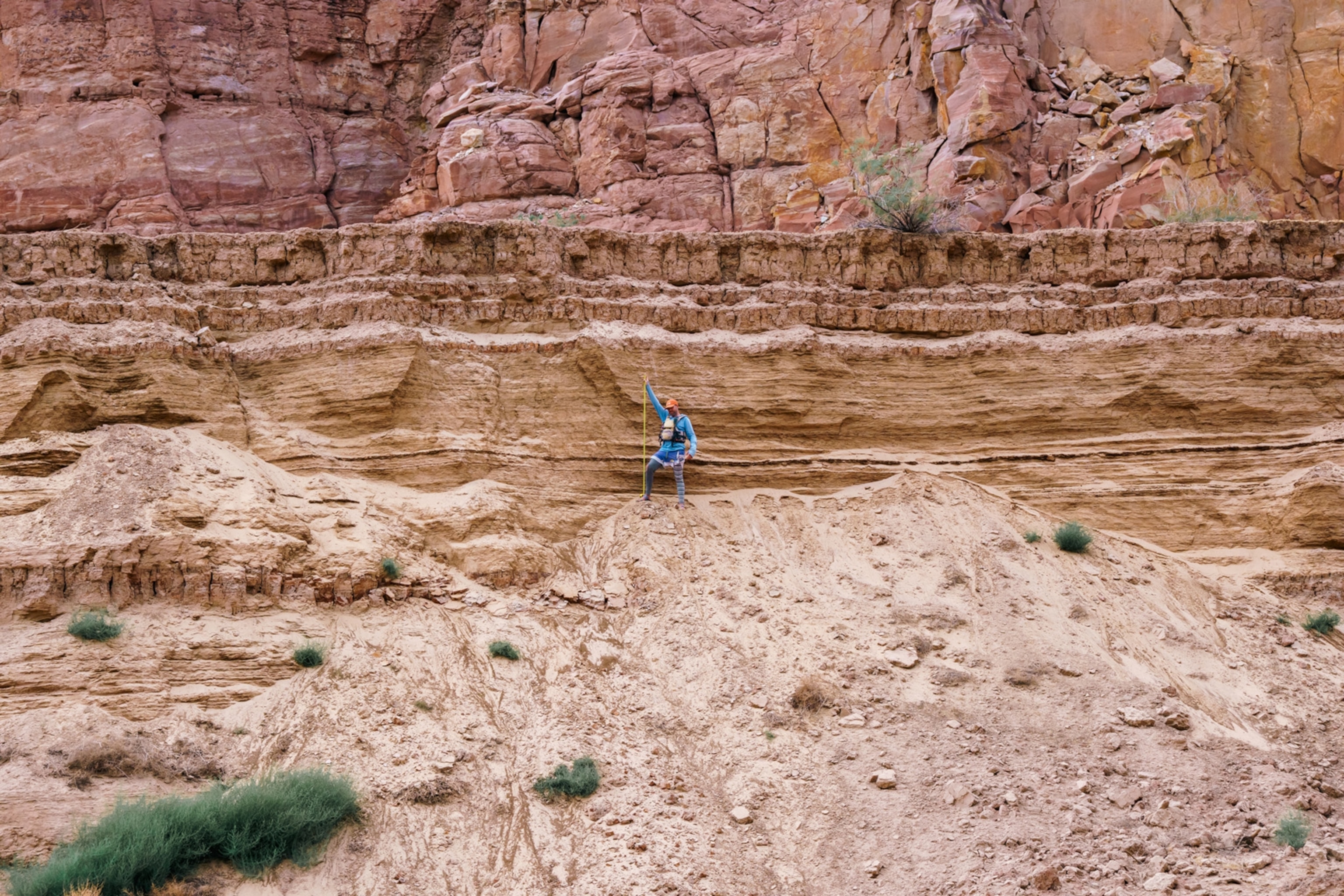 A person wearing a blue shirt and an orange baseball cap holds a yellow stick in front of a wall of sediment.