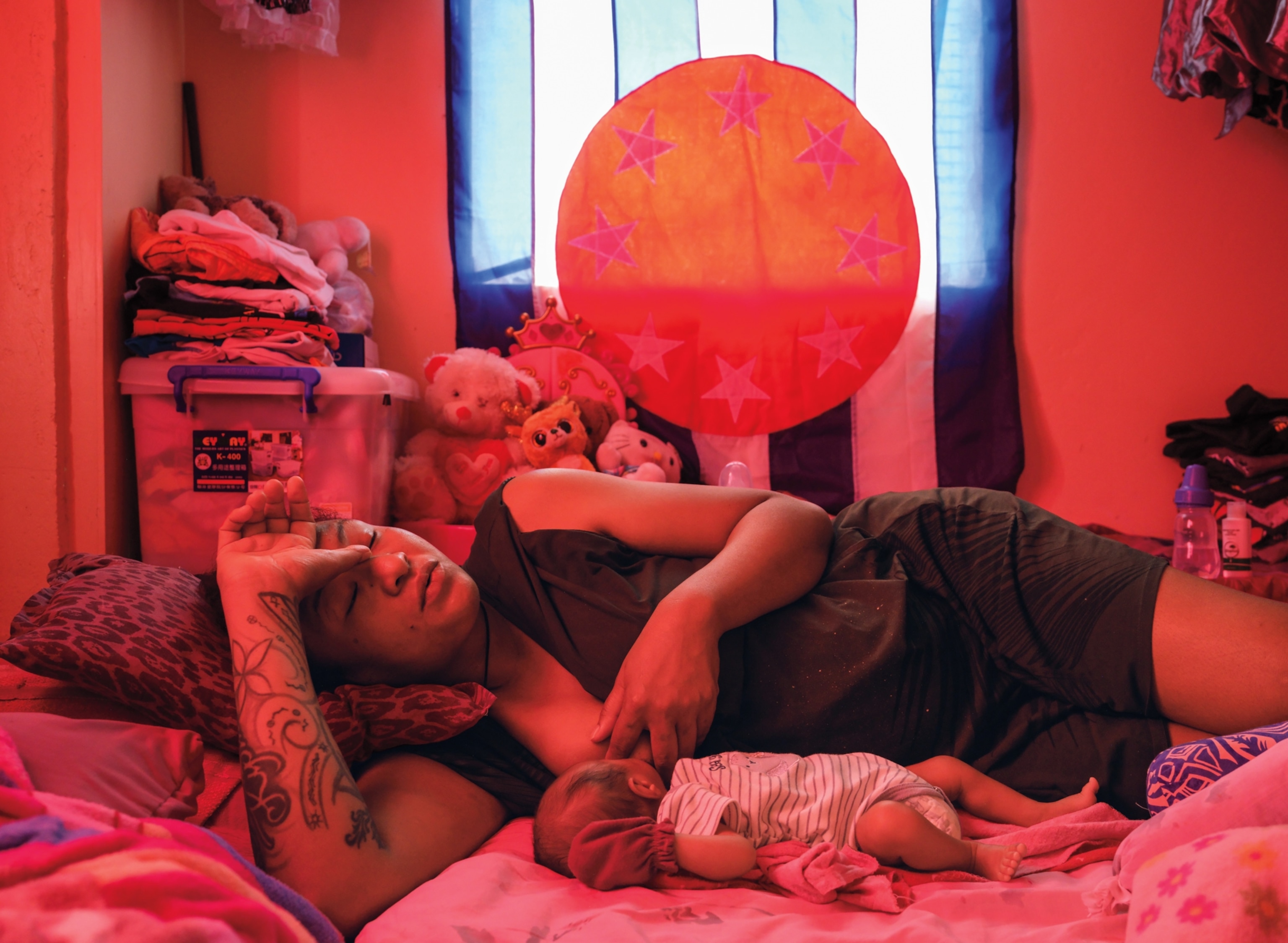A woman lays on her side on a bed while her baby nurses from her breast. In the background are various clothes and stuffed animals and the room glows slightly red.
