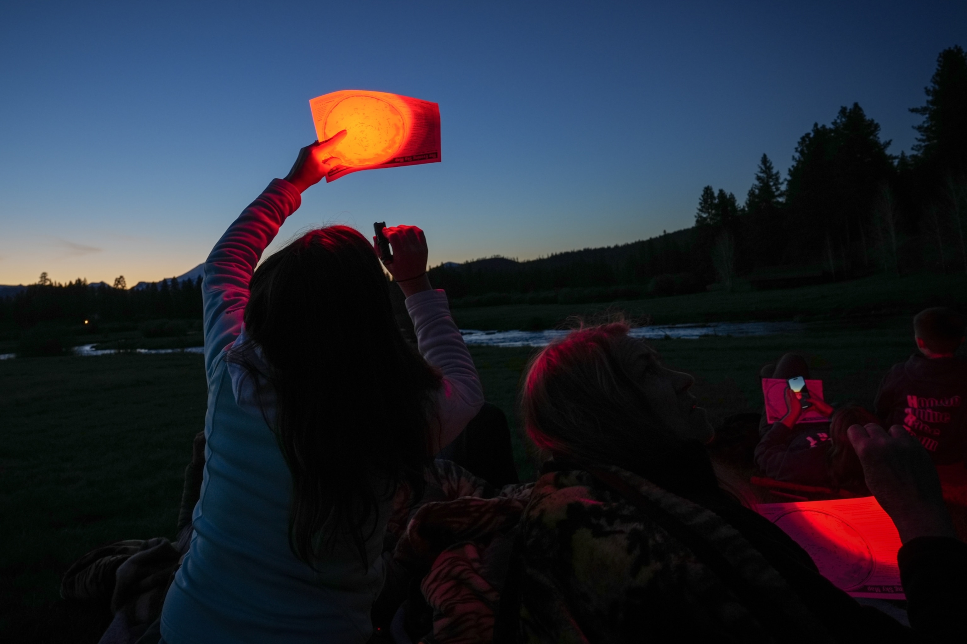 People are gathered at dusk, with a woman shining a red light onto a sky chart as she looks up at the sky above